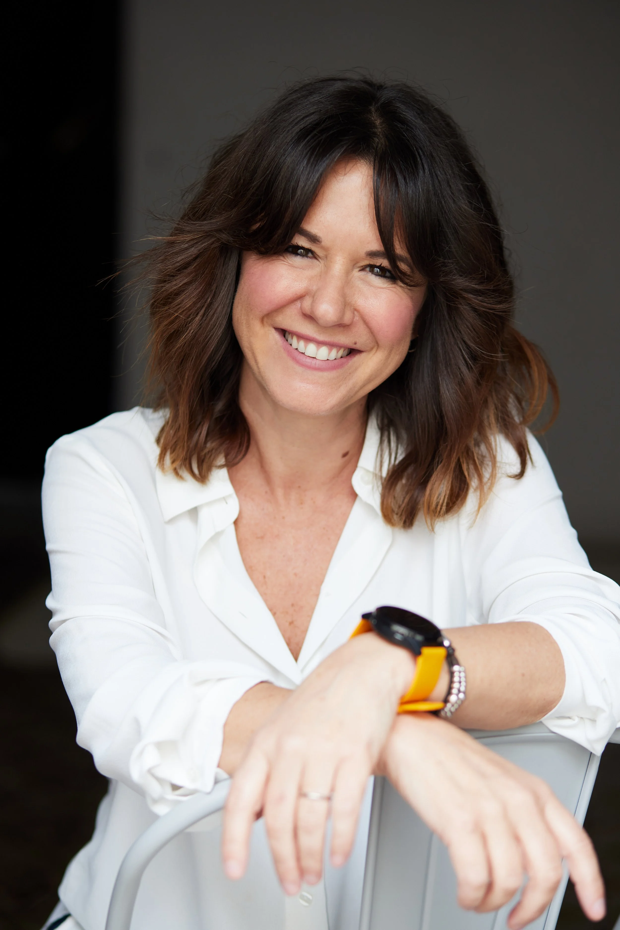 A woman with shoulder-length brown hair smiling, wearing a white shirt, sitting at a table with her arms resting on the back of a chair, and wearing a yellow and black wristwatch.