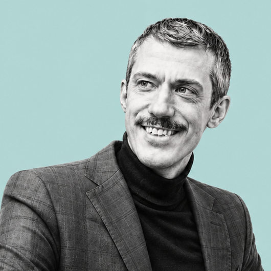 Black and white portrait of a smiling man with a mustache, wearing a dark turtleneck and a blazer, against a light blue background.