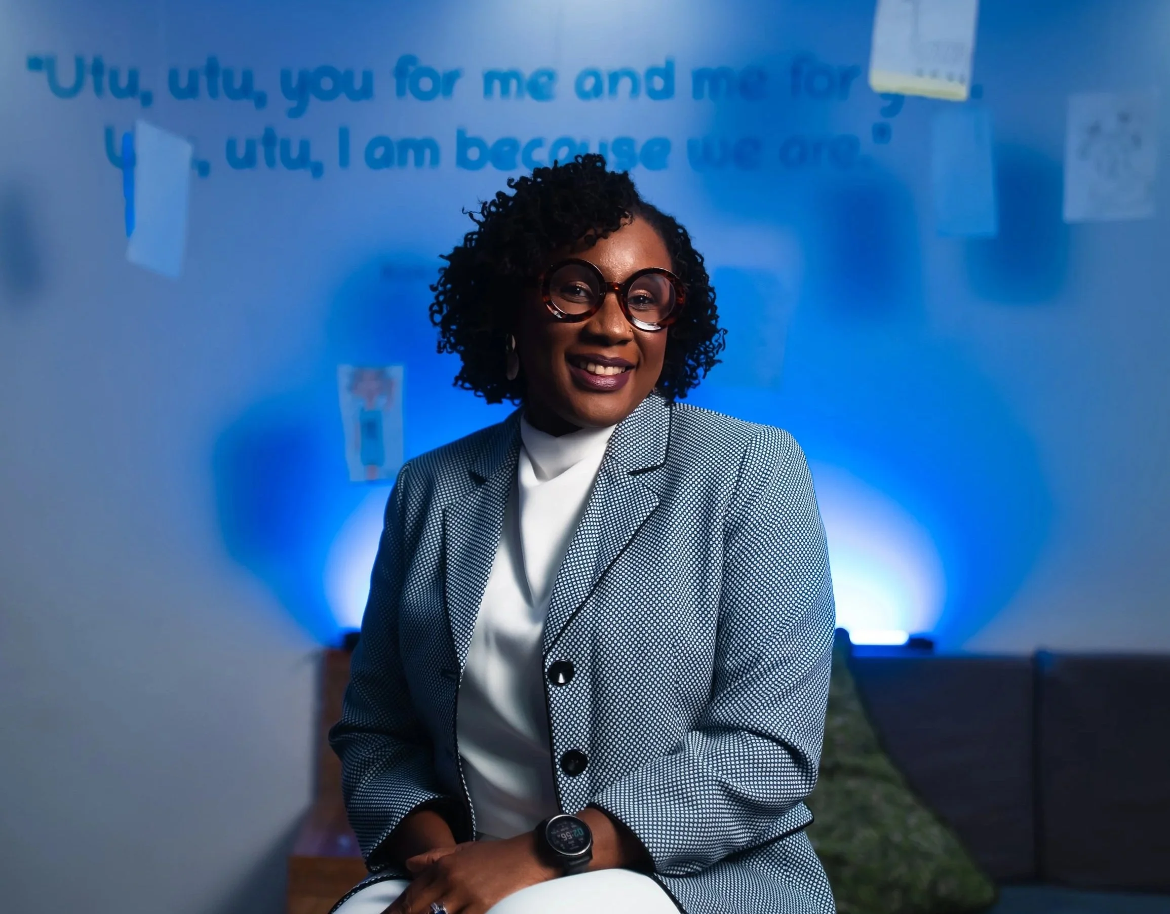 A woman with curly hair, wearing glasses, a checkered blazer, and a smartwatch, smiling in front of a blue wall with a quote written on it.