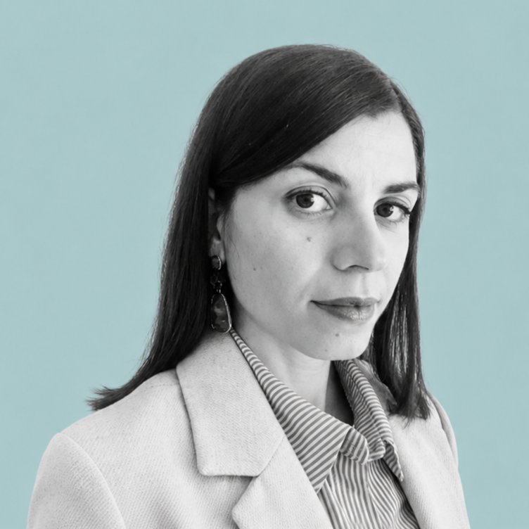 A woman with dark hair and earrings looking at the camera, wearing a light-colored blazer over a striped shirt with a solid background.