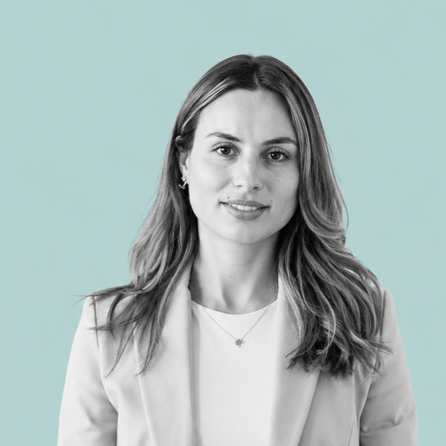 Portrait of a young woman with shoulder-length wavy hair, wearing a light blazer and a necklace, against a light blue background.
