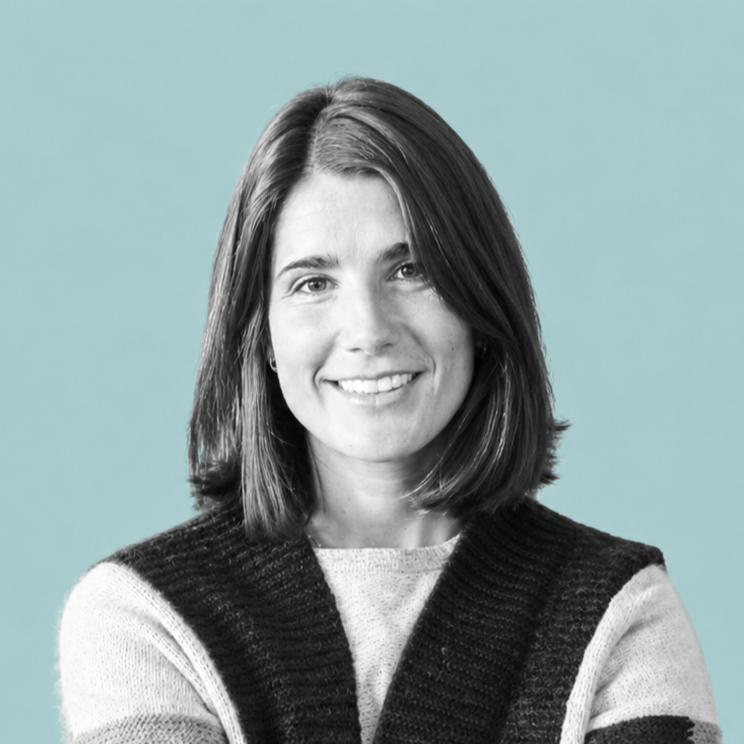 A woman with shoulder-length brown hair, smiling, wearing a black and white knitted sweater, against a light blue background.