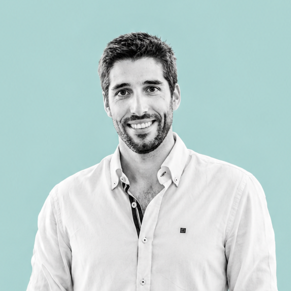 A man with dark hair and a beard smiling, wearing a light-colored button-up shirt with a collar, against a turquoise background.