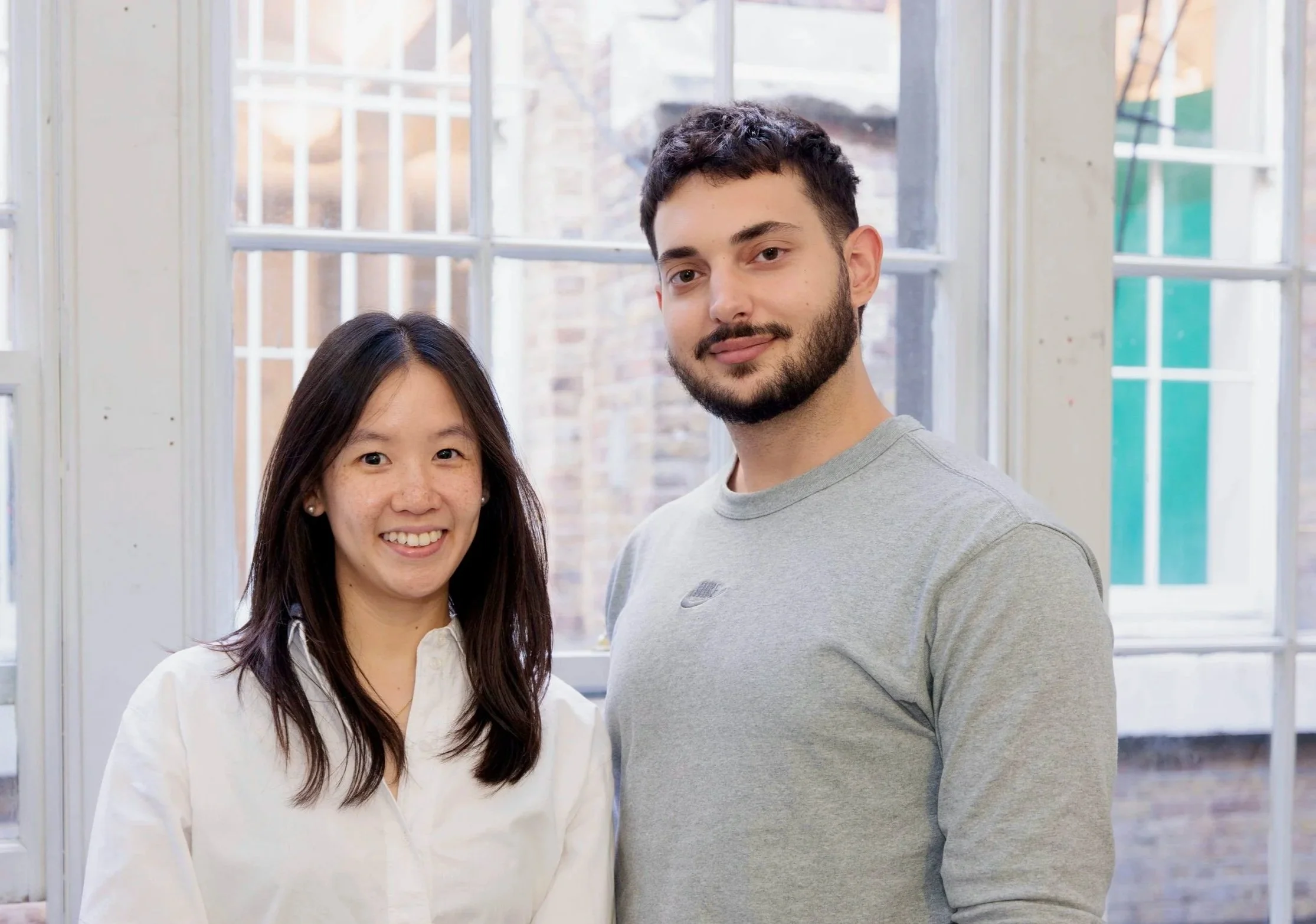 A young Asian woman with long dark hair and a young Caucasian man with short dark hair and a beard standing inside a room in front of a large window.