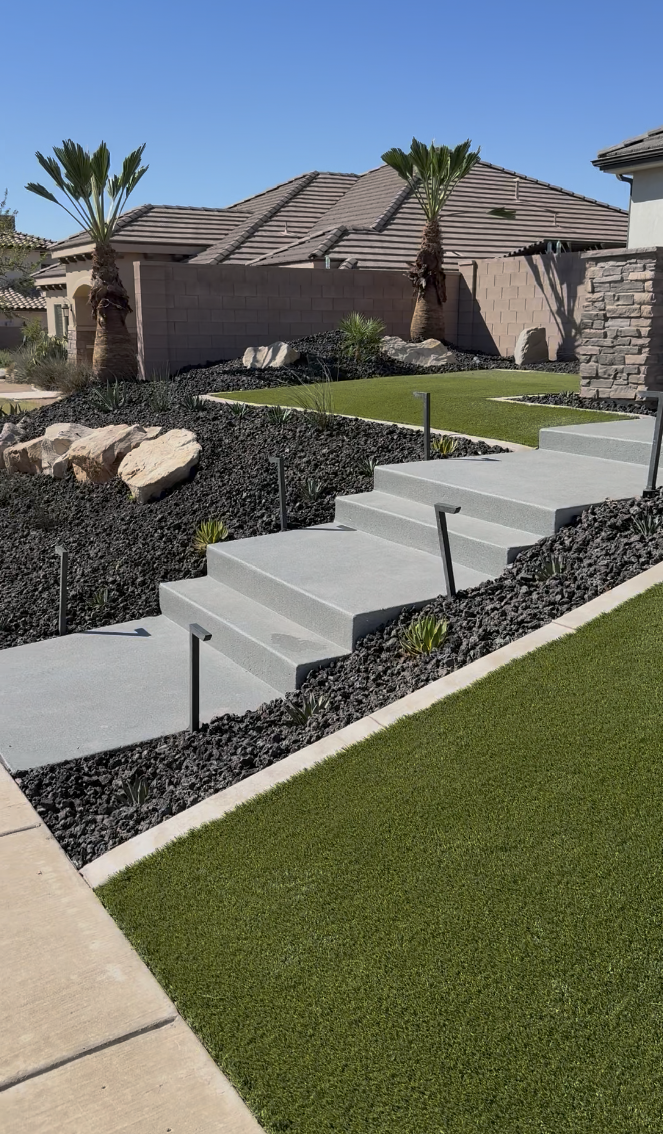A modern backyard with a concrete staircase leading to a landscaped yard with green grass and desert plants, including two palm trees, rocks, and black gravel, with a brick wall and house with tiled roof in the background.