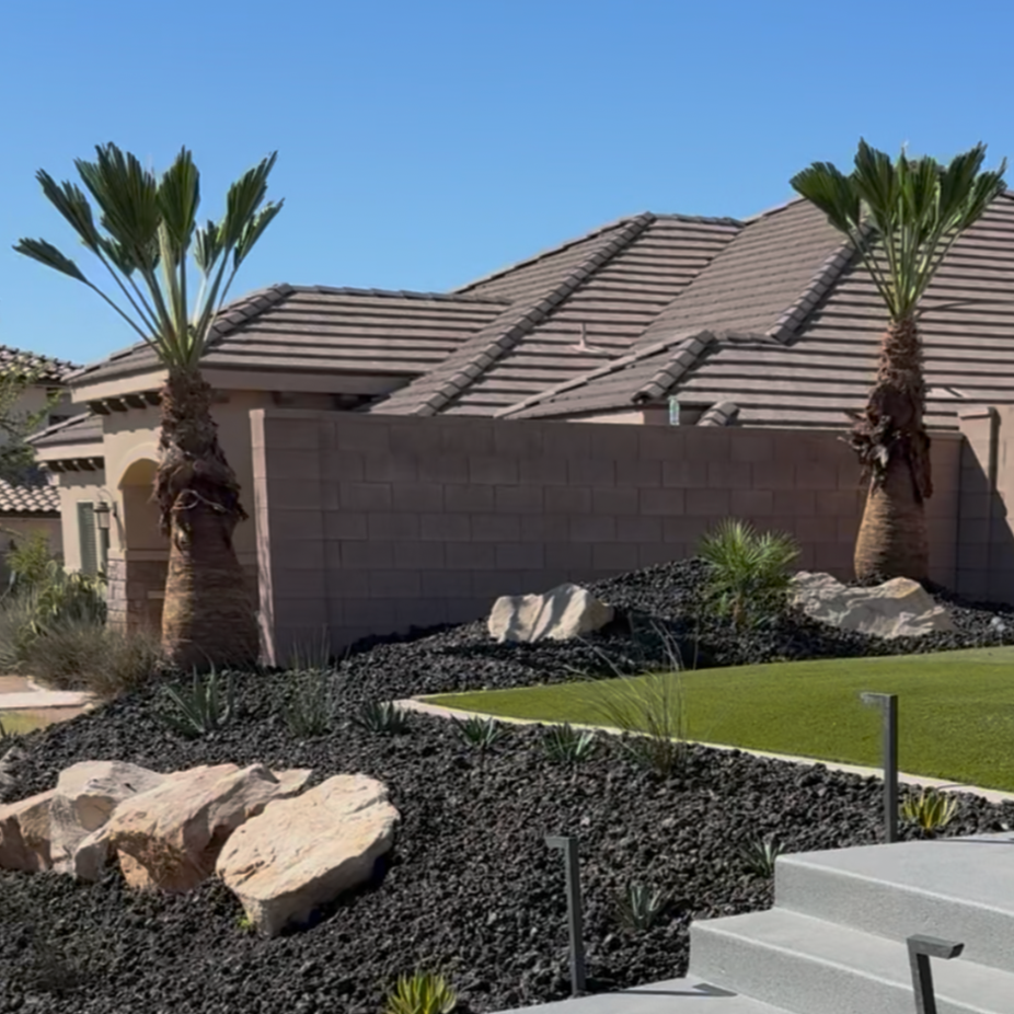 Front yard of a house featuring two palm trees, large rocks, desert plants, a manicured lawn, and steps leading up to the porch with a clear blue sky overhead.