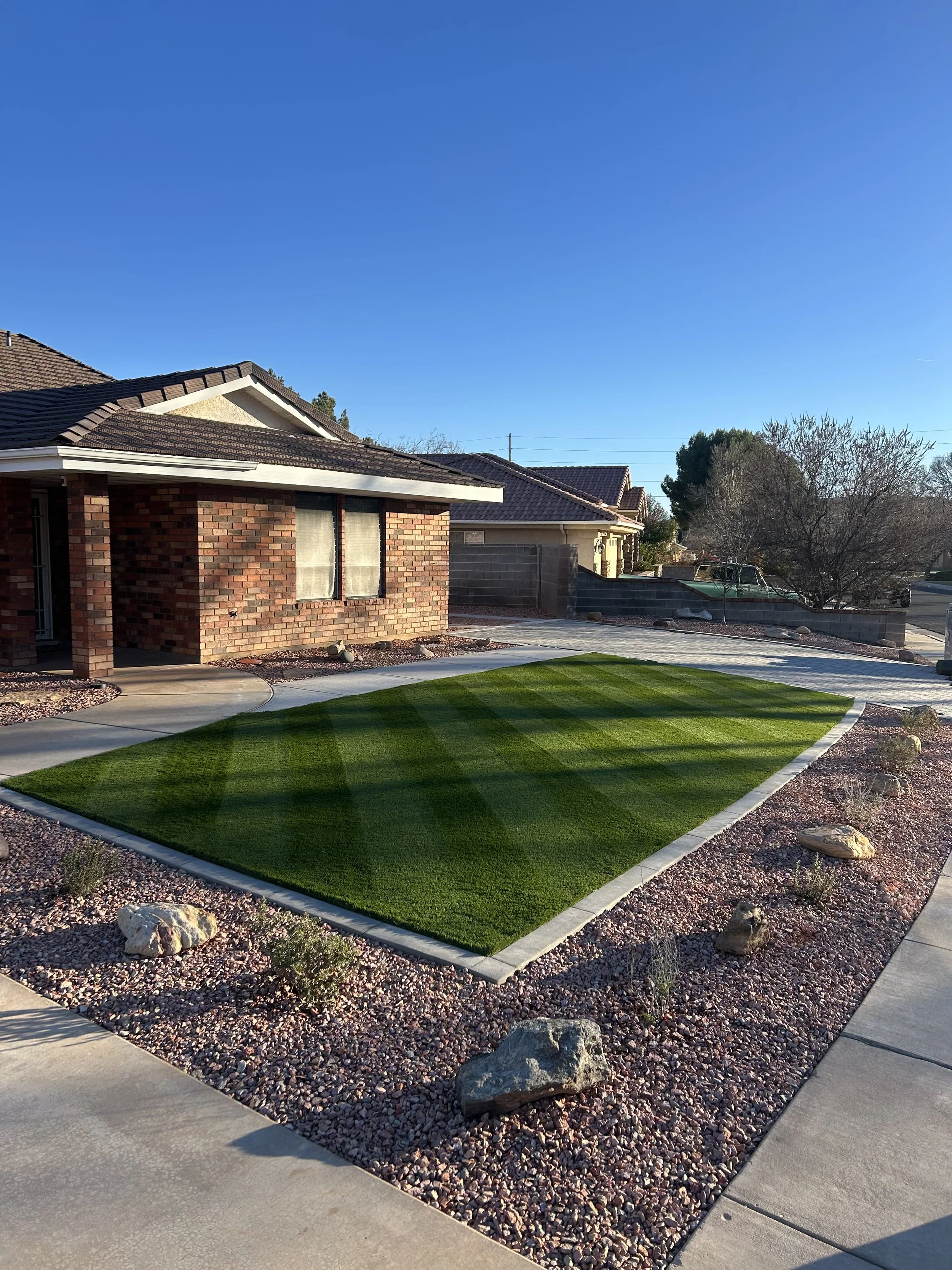 A front yard with a lawn, shrubbery, and a concrete walkway. There is a brick house with a dark shingled roof on the left side of the image. The sky is clear and blue, and there are trees and neighboring houses in the background.