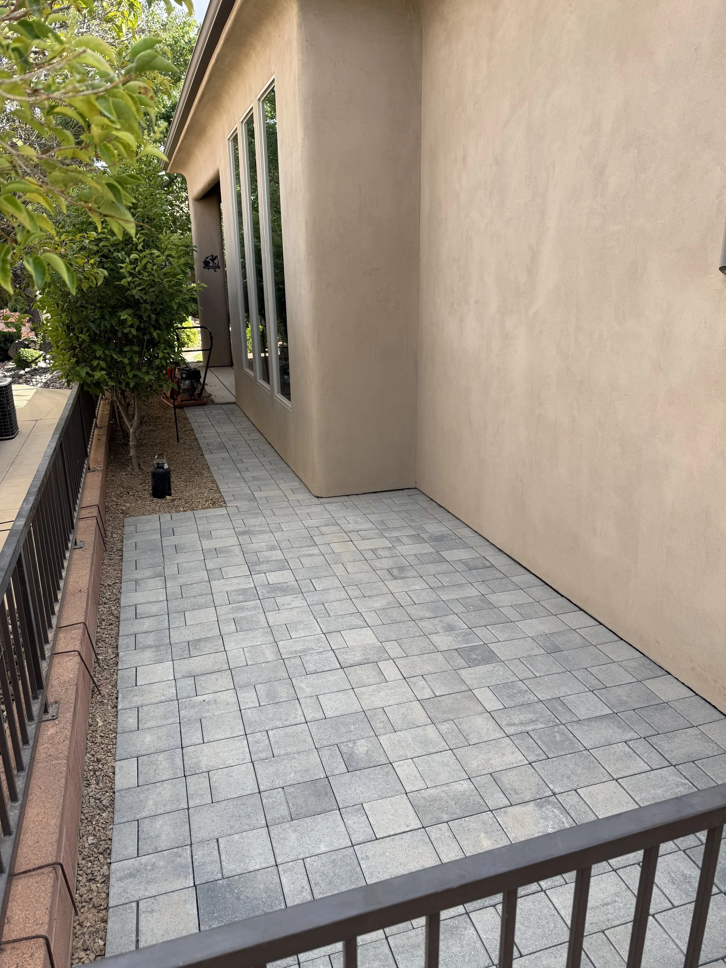 View of a small backyard patio enclosed by a black metal railing, with a pathway made of gray paver stones, next to a beige stucco house wall with three tall windows, a tree with green leaves, and some gardening equipment.