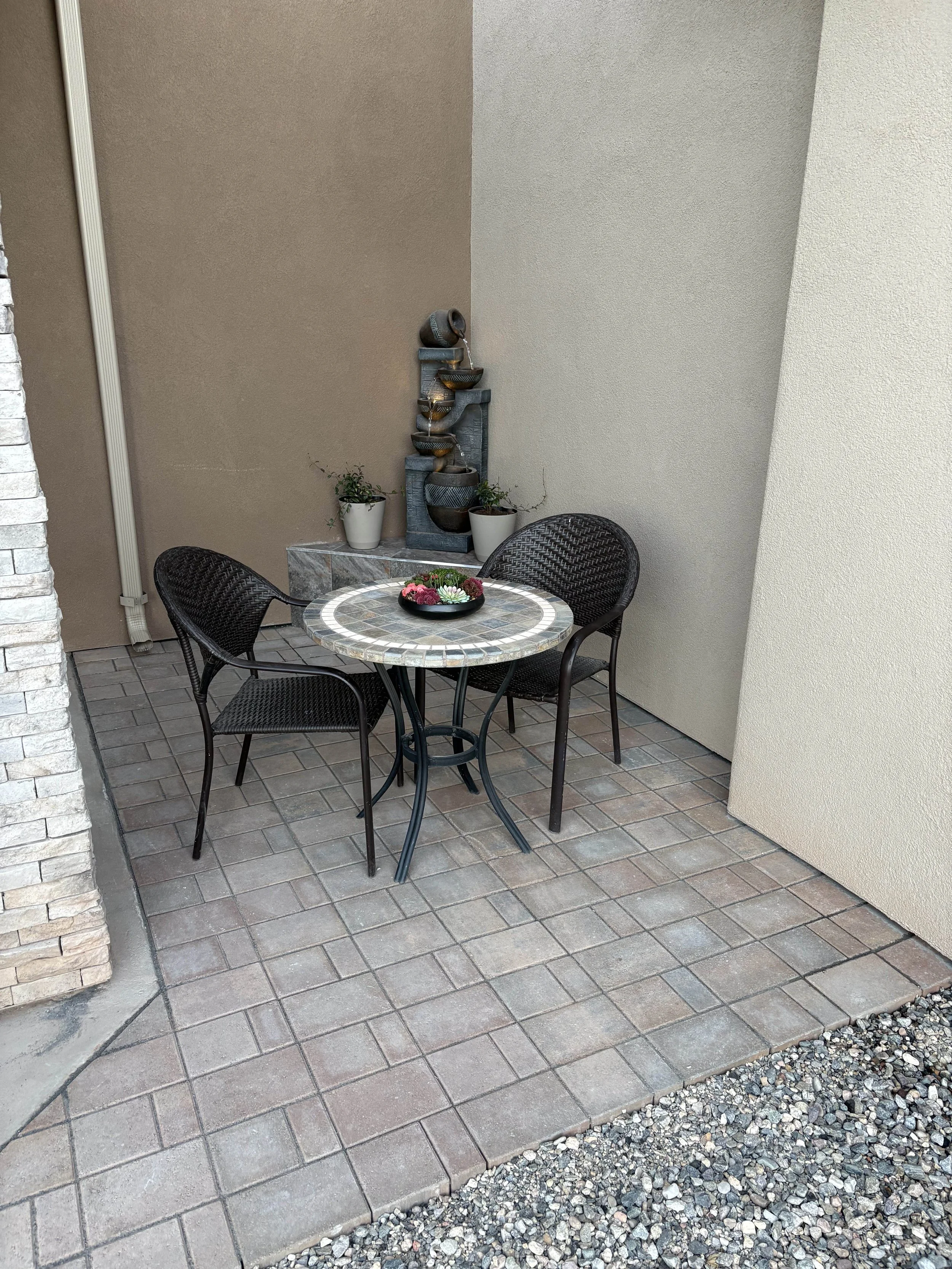 An outdoor patio space with a round mosaic table and four black wicker chairs. There is a small water fountain with three bowls and potted plants against a beige and light gray wall. The patio has brick pavers and a gravel border.