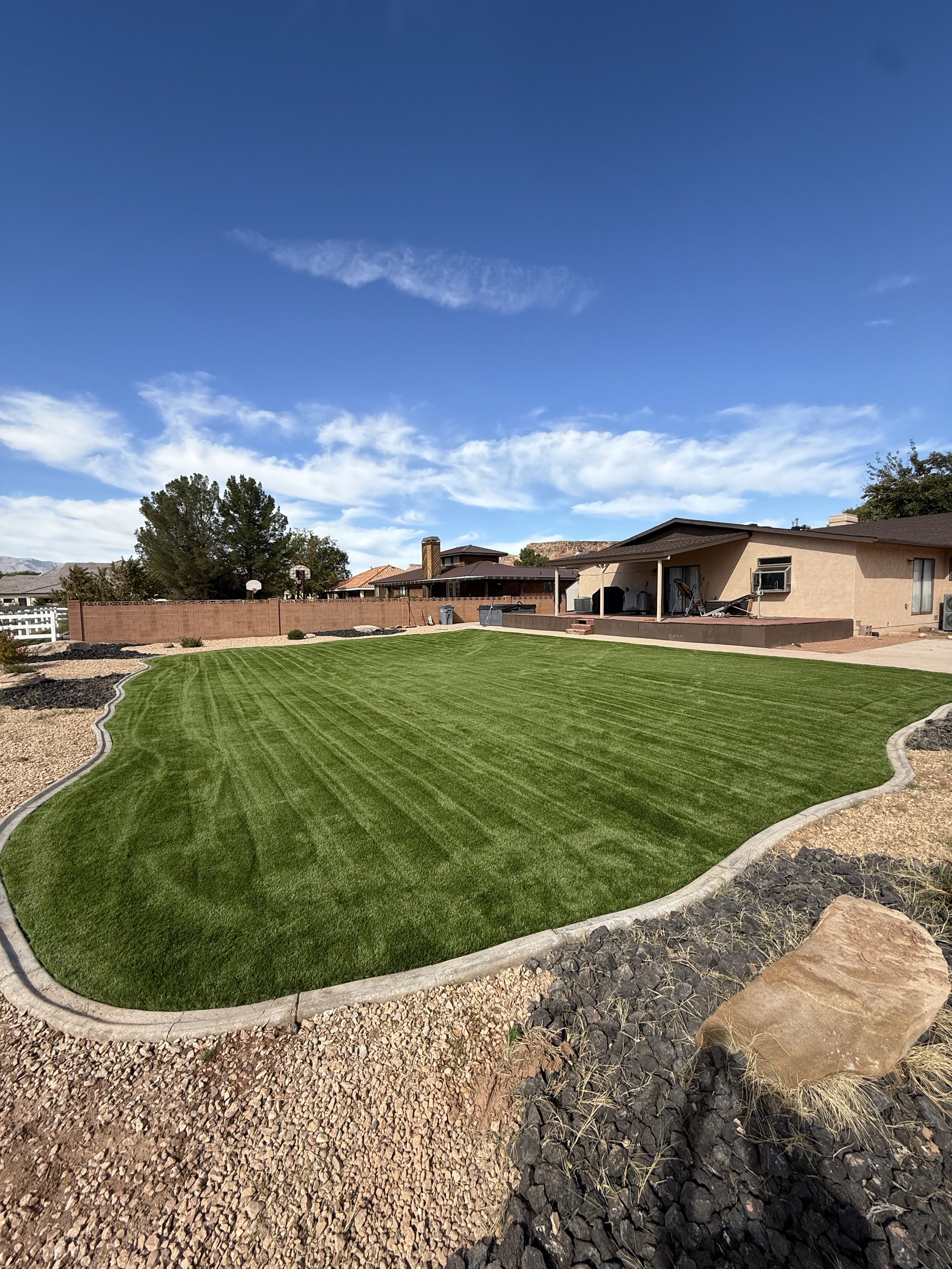 Backyard with a well-maintained green lawn, a beige house with a covered patio, a brick wall fence, trees, and a partly cloudy blue sky.
