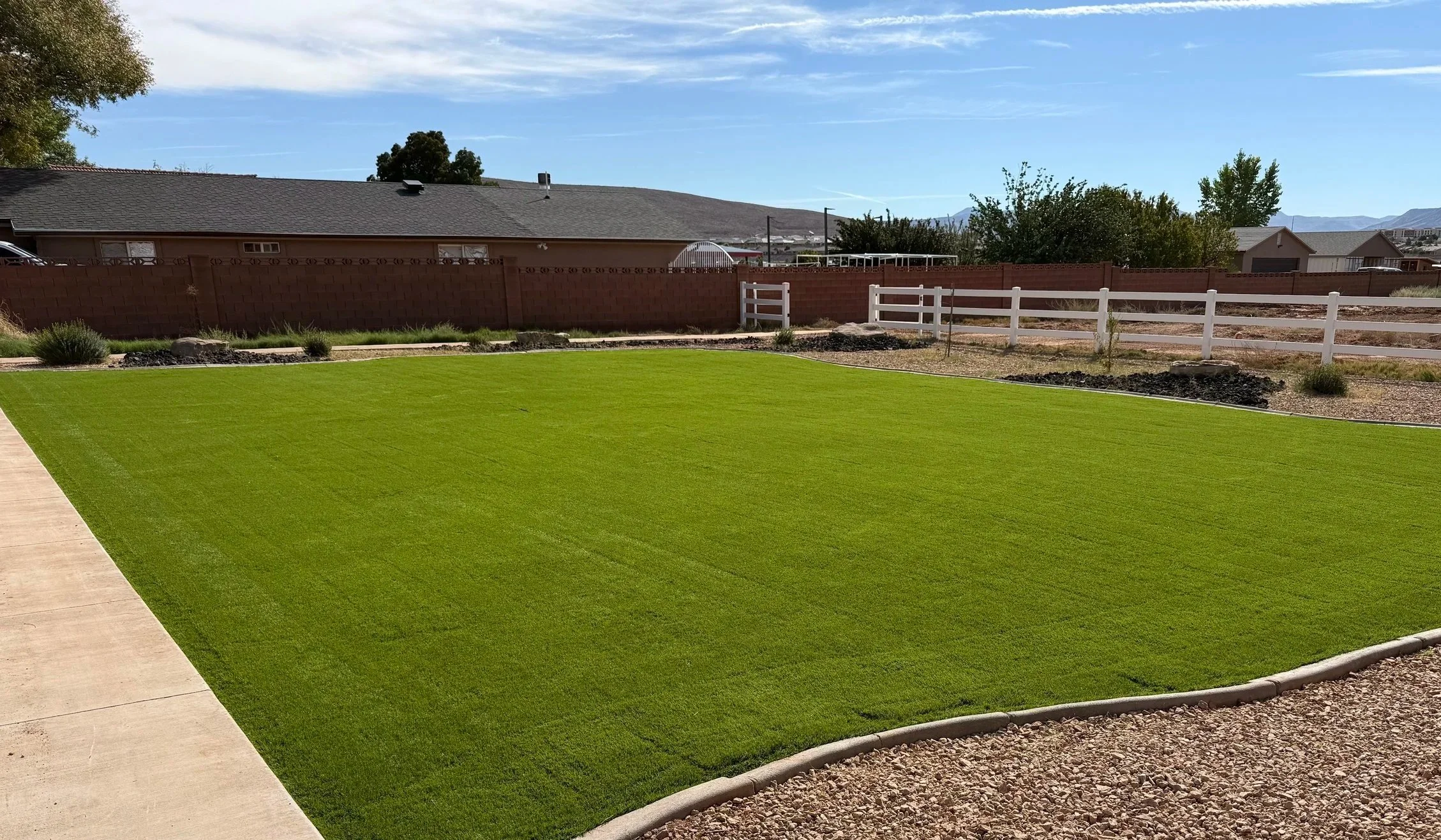 A backyard with a well-maintained green lawn, a concrete pathway on the left, surrounded by a red brick fence and a white wooden fence, with trees, houses, and mountains visible in the background under a partly cloudy sky.
