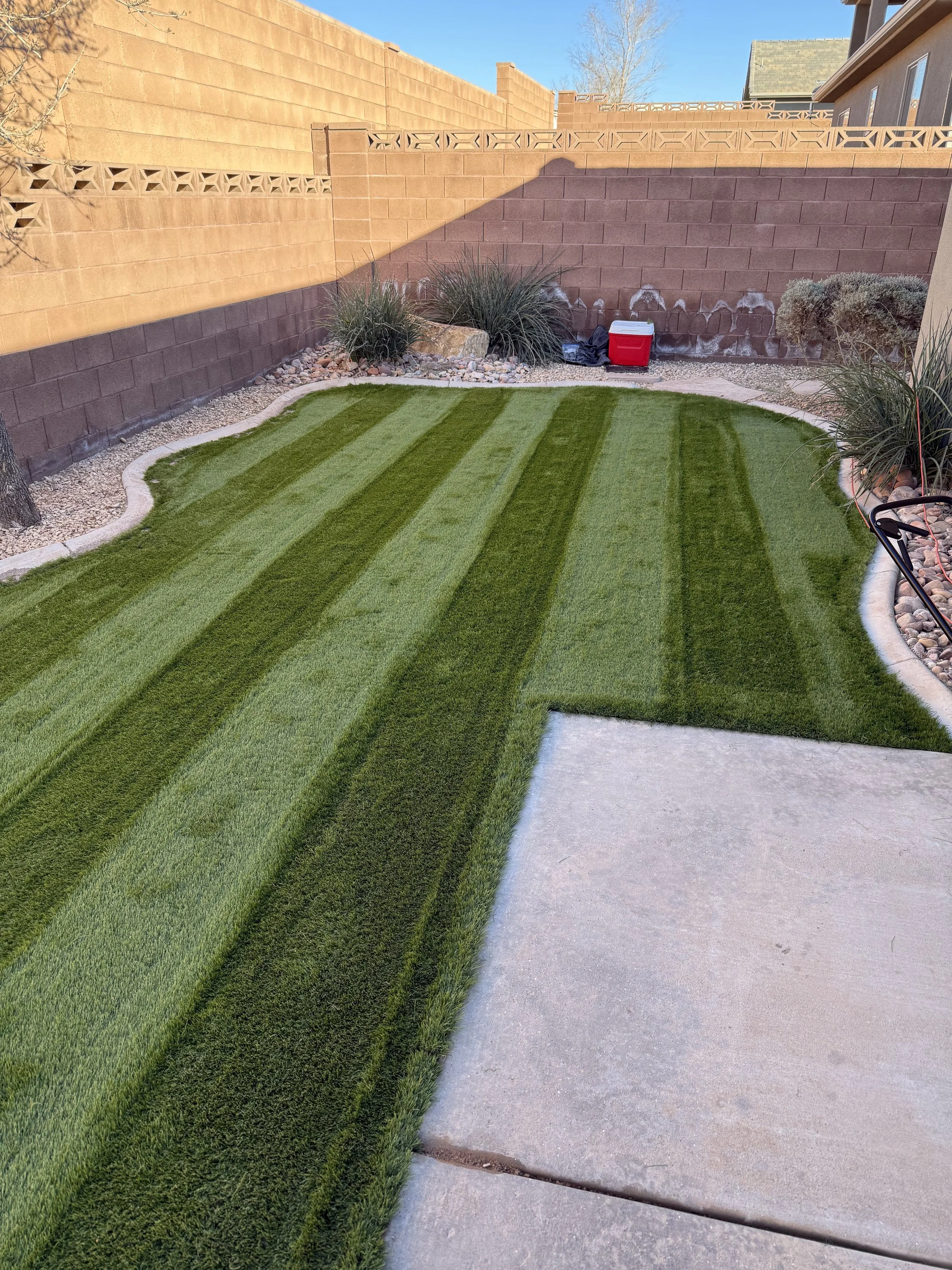 Backyard with artificial grass, plants, a brick wall, and a cooler.