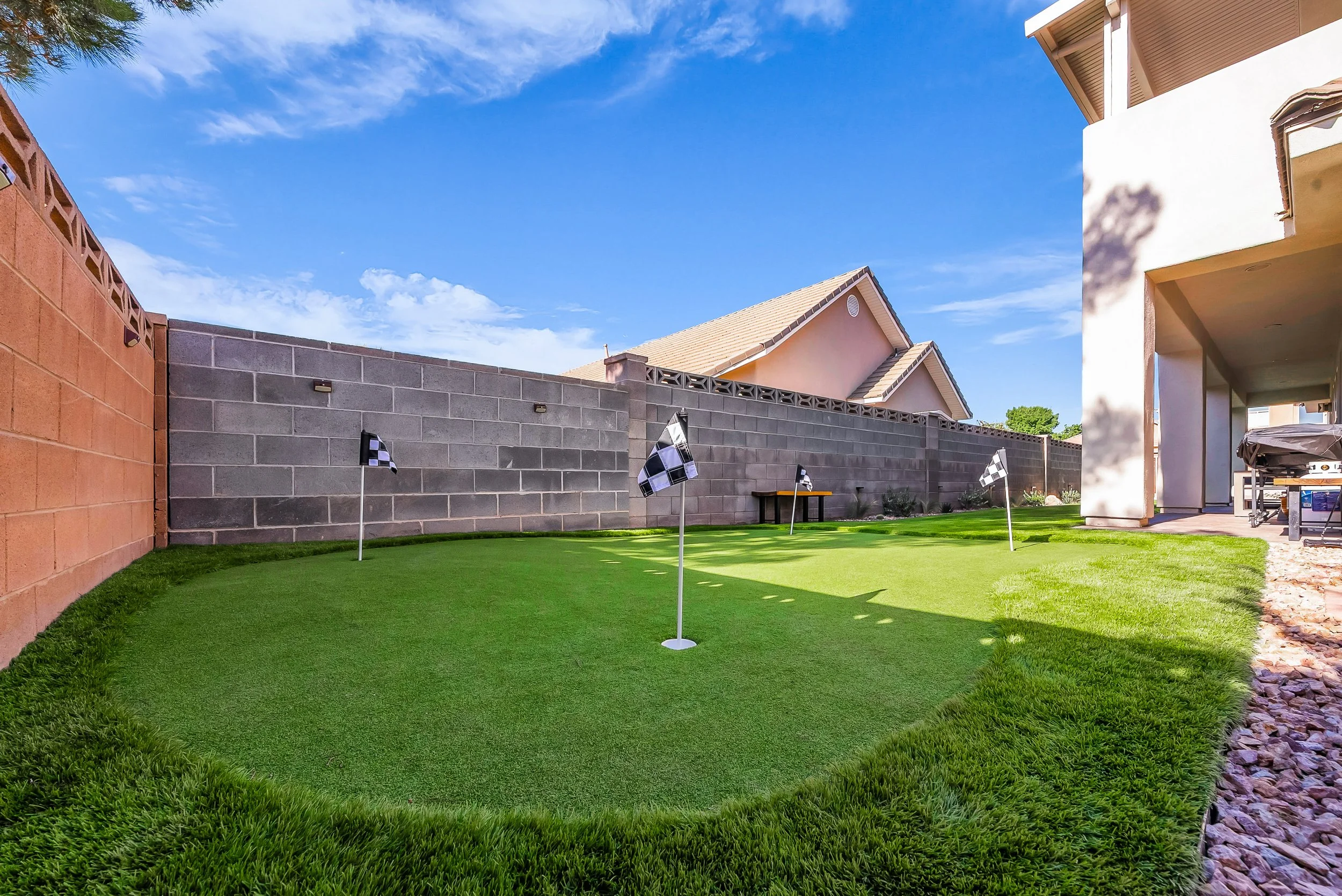 Backyard with a green grass lawn, a white house with patio furniture, a small slide, and a garden bed with plants, enclosed by a concrete block wall, under a blue sky with clouds.