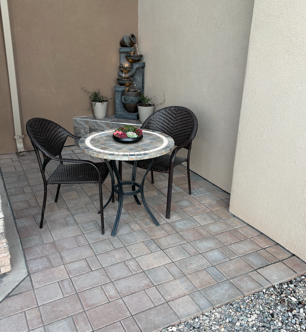 Outdoor patio with a round table and two wicker chairs, a small wall fountain, and potted plants in the background.