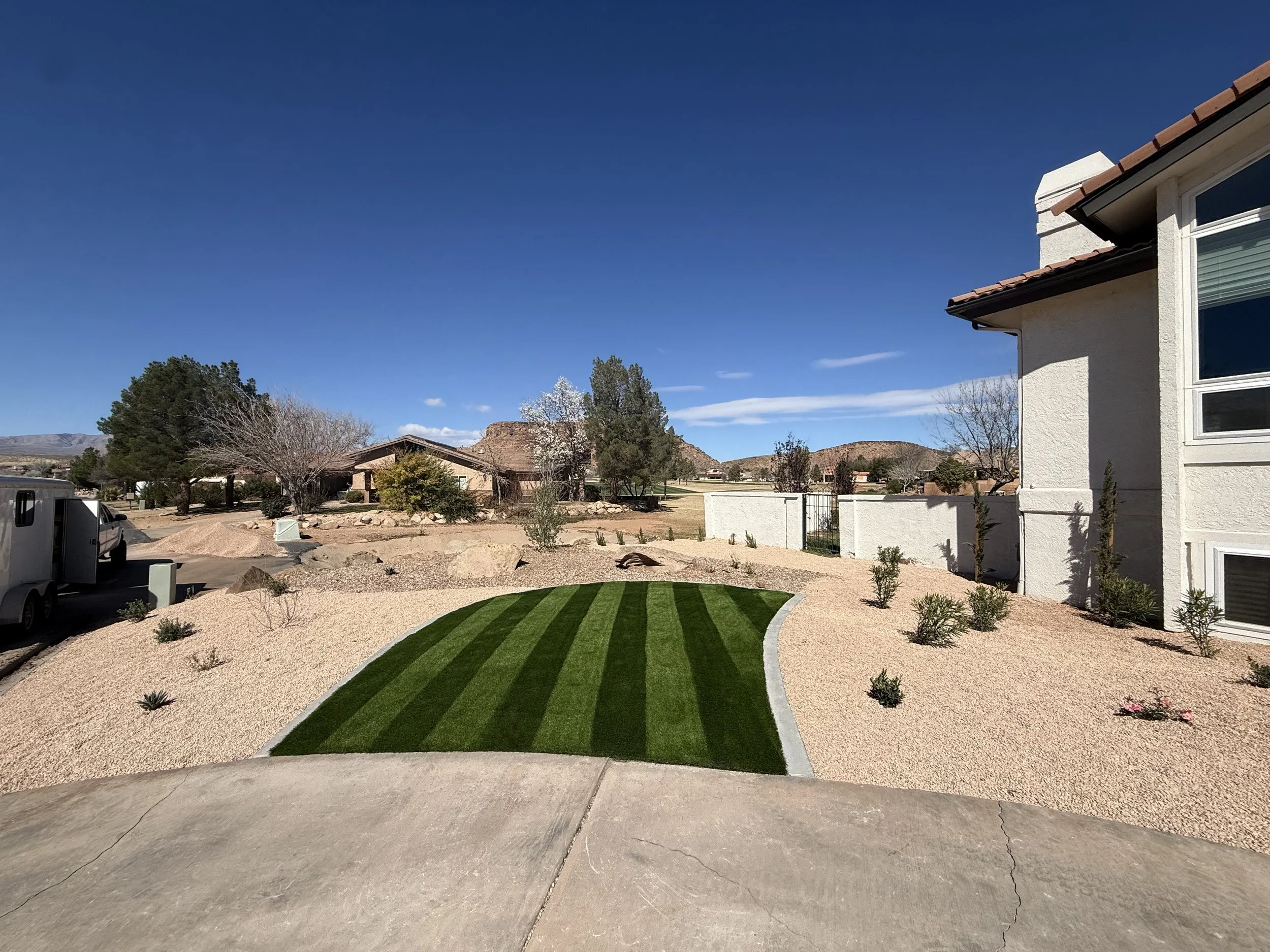 A residential backyard with a curved patch of freshly laid green grass, surrounded by light-colored gravel and small plants. A two-story house with a white exterior and large windows is visible on the right. In the background, there are trees, other 