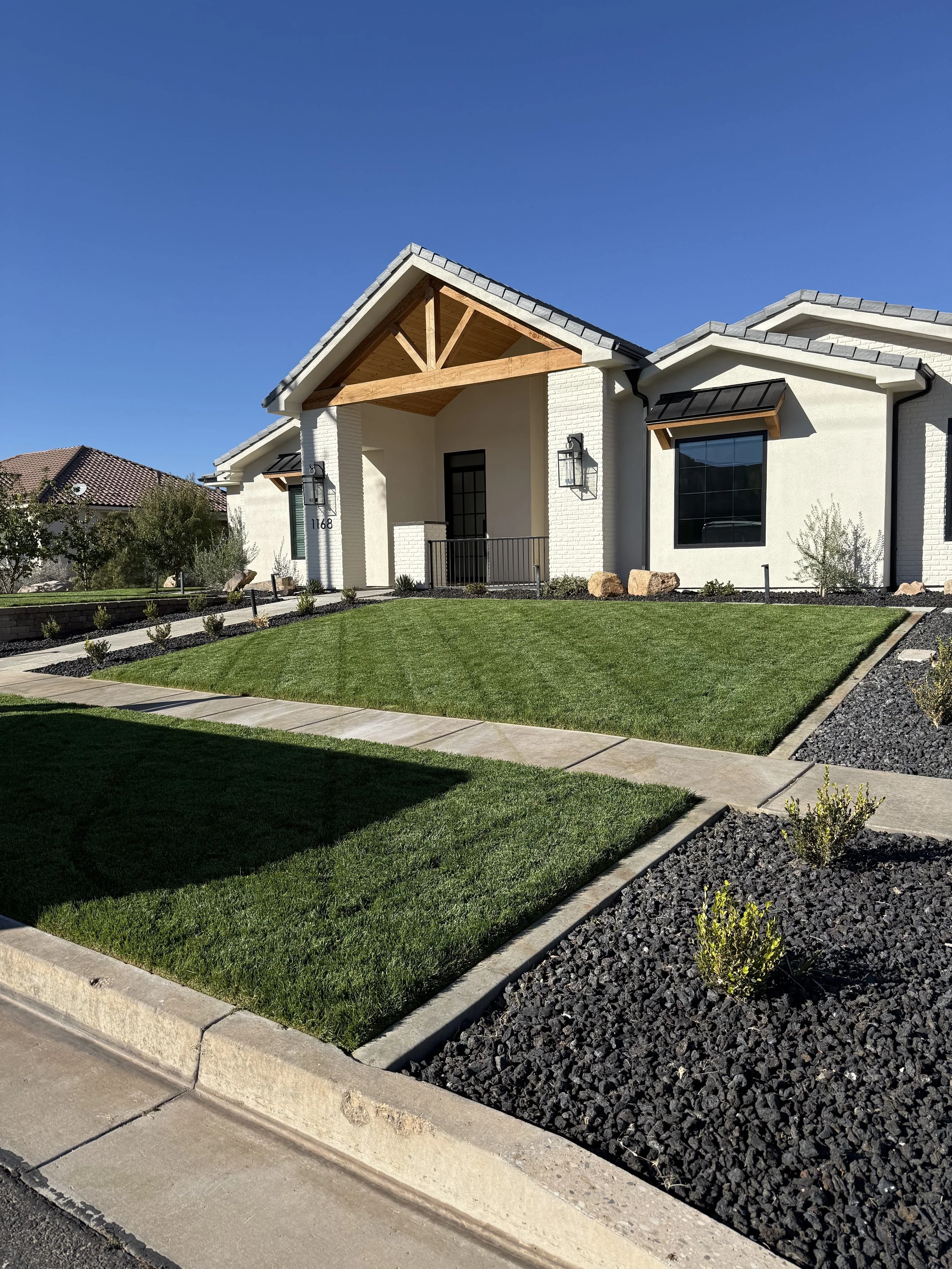 A modern white house with a gray shingle roof, front porch, and a manicured lawn with landscaped bushes and rocks.