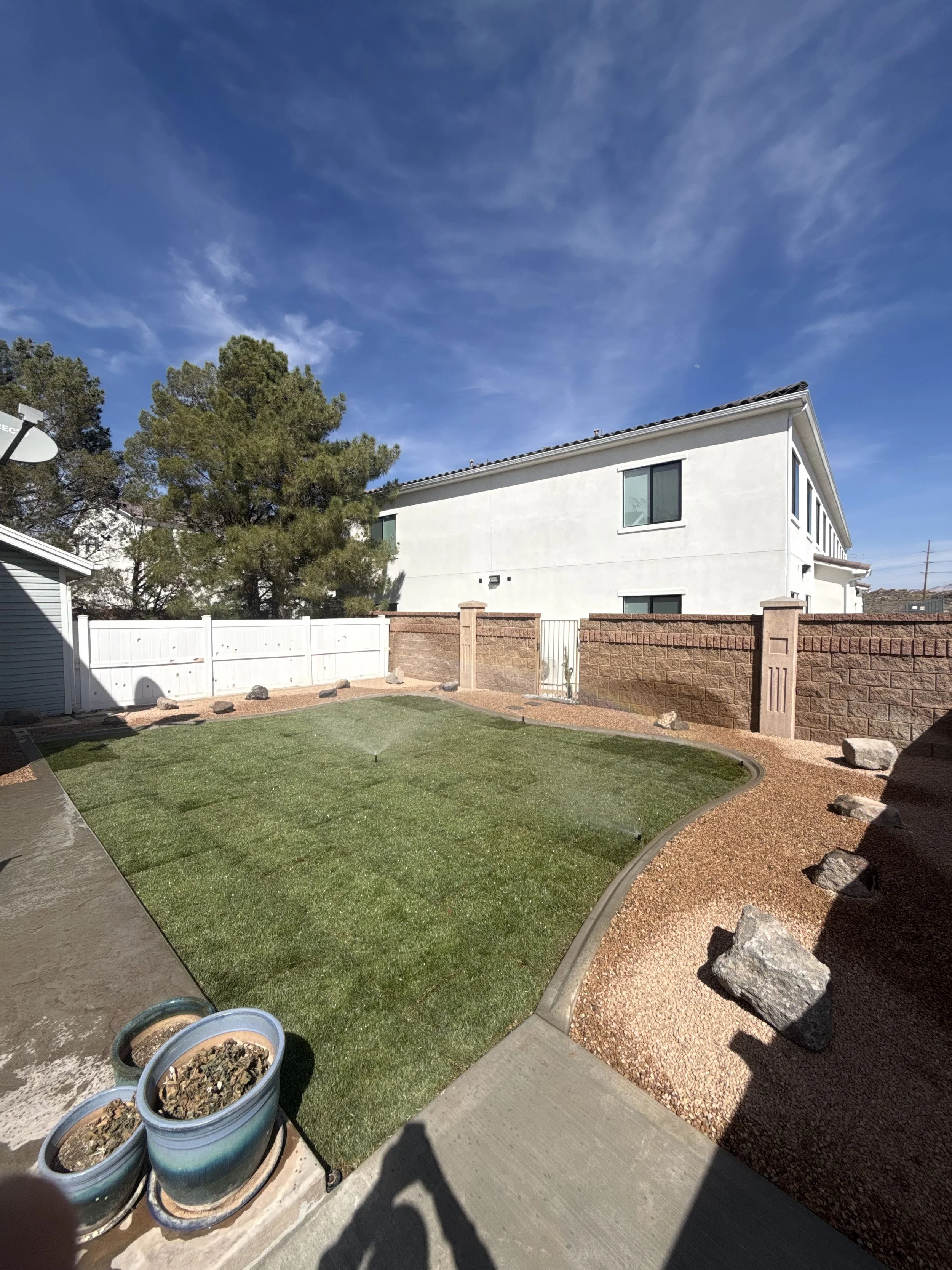 A backyard with a small patch of artificial grass, a curved brick border, a red brick wall, and a white house with several windows. There are large rocks along the border and a few buckets filled with dirt or debris. The sky is blue with some clouds,