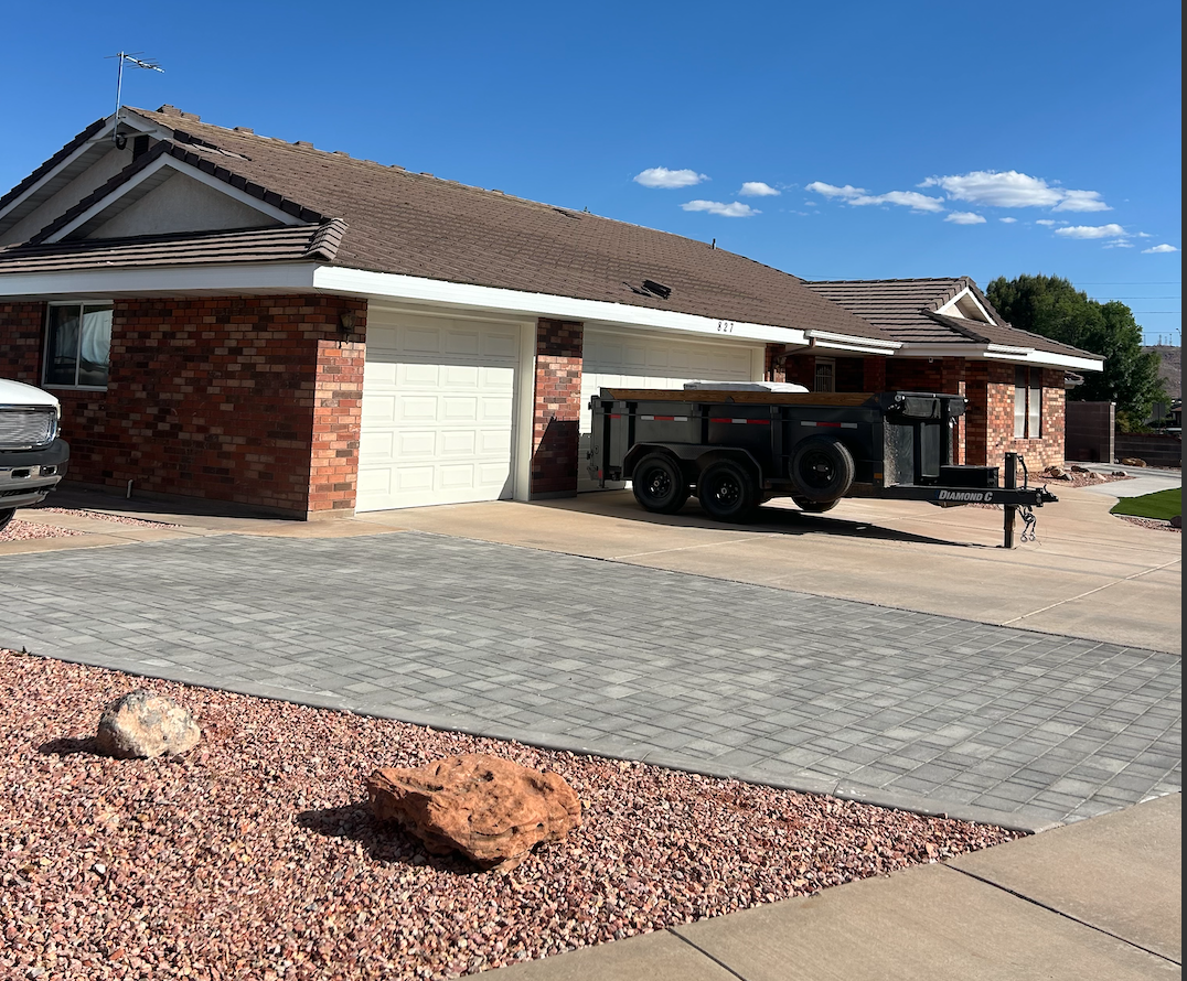 A brick house with a brown tiled roof and a white garage door. There is a black trailer parked on the driveway and a vehicle partially visible on the left. The landscaping includes rocks and gravel, with a concrete driveway and patio under a blue sky with some clouds.