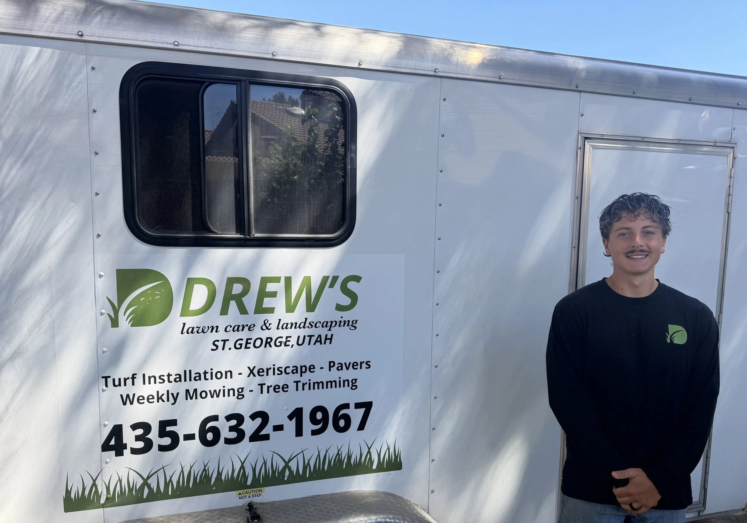 A person stands outside a white trailer with green and black text advertising lawn care and landscaping services in St. George, Utah. The trailer has a window and a door, with a reflection of houses and trees visible in the window.