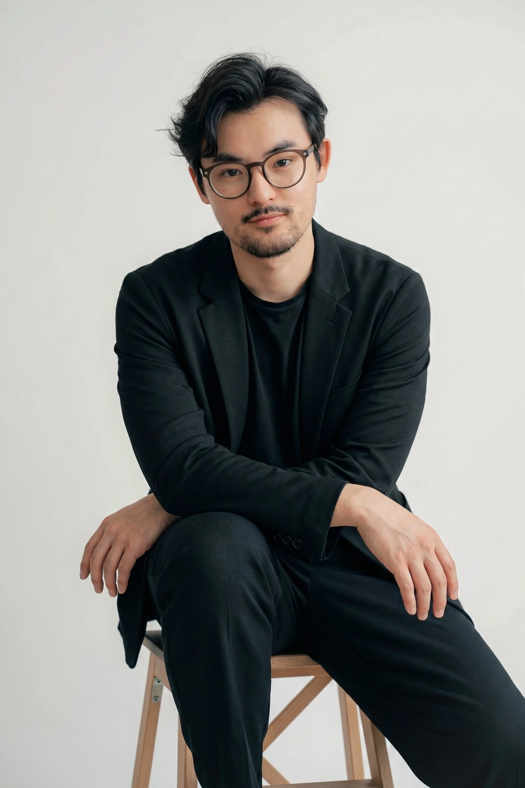 A young man with black hair and glasses sitting on a wooden stool against a plain white background.