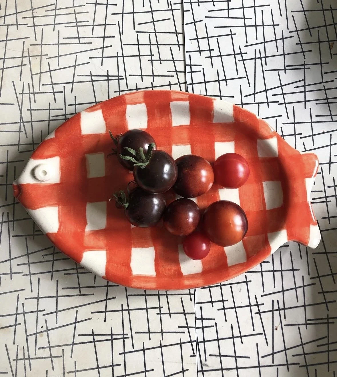 A fish-shaped dish with a red and white checkered pattern, containing a handful of dark and red grapes, placed on a surface with black intersecting lines.