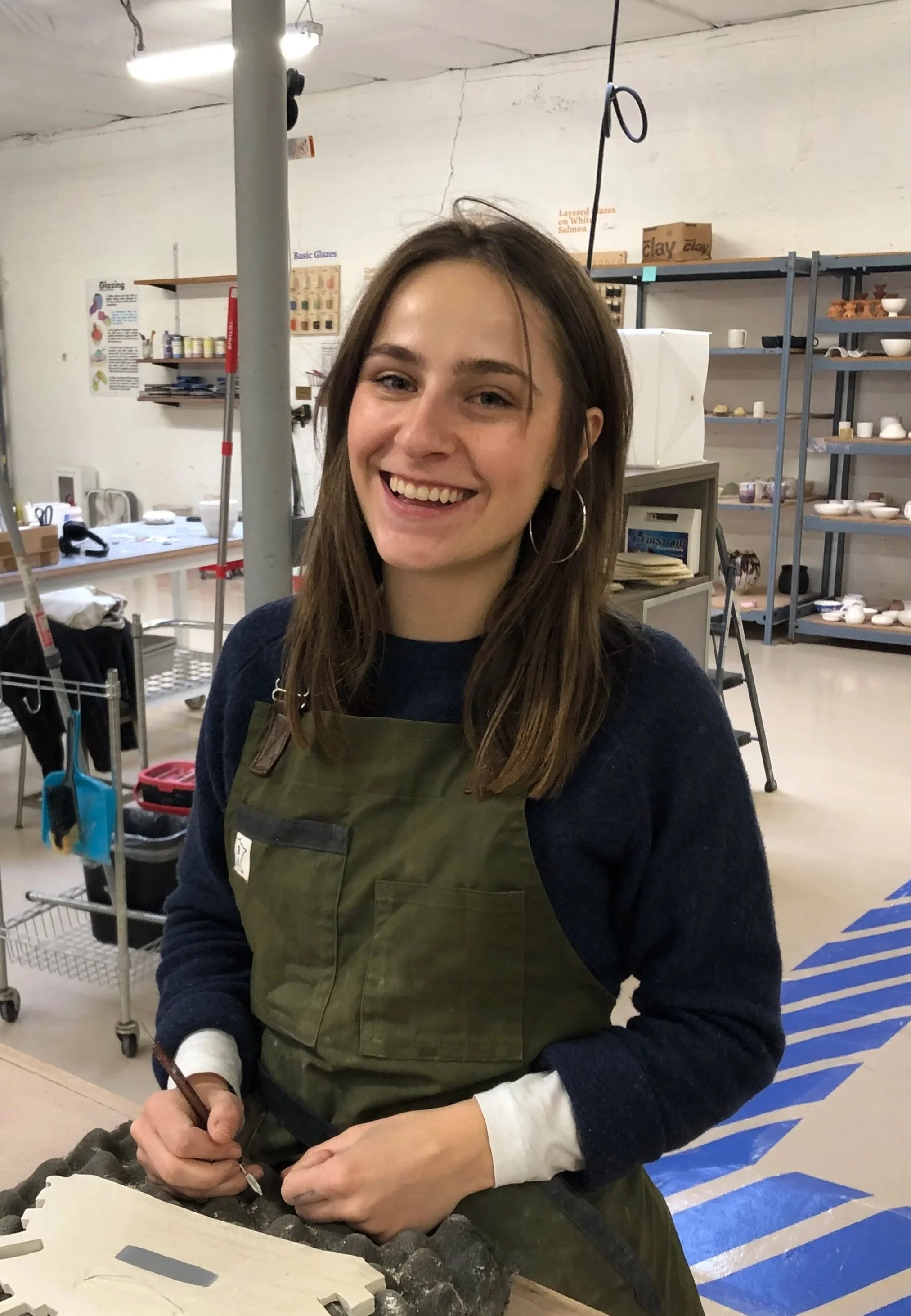 Young woman smiling and working with a tool in a ceramics or pottery studio, surrounded by shelves with bowls and pottery items.