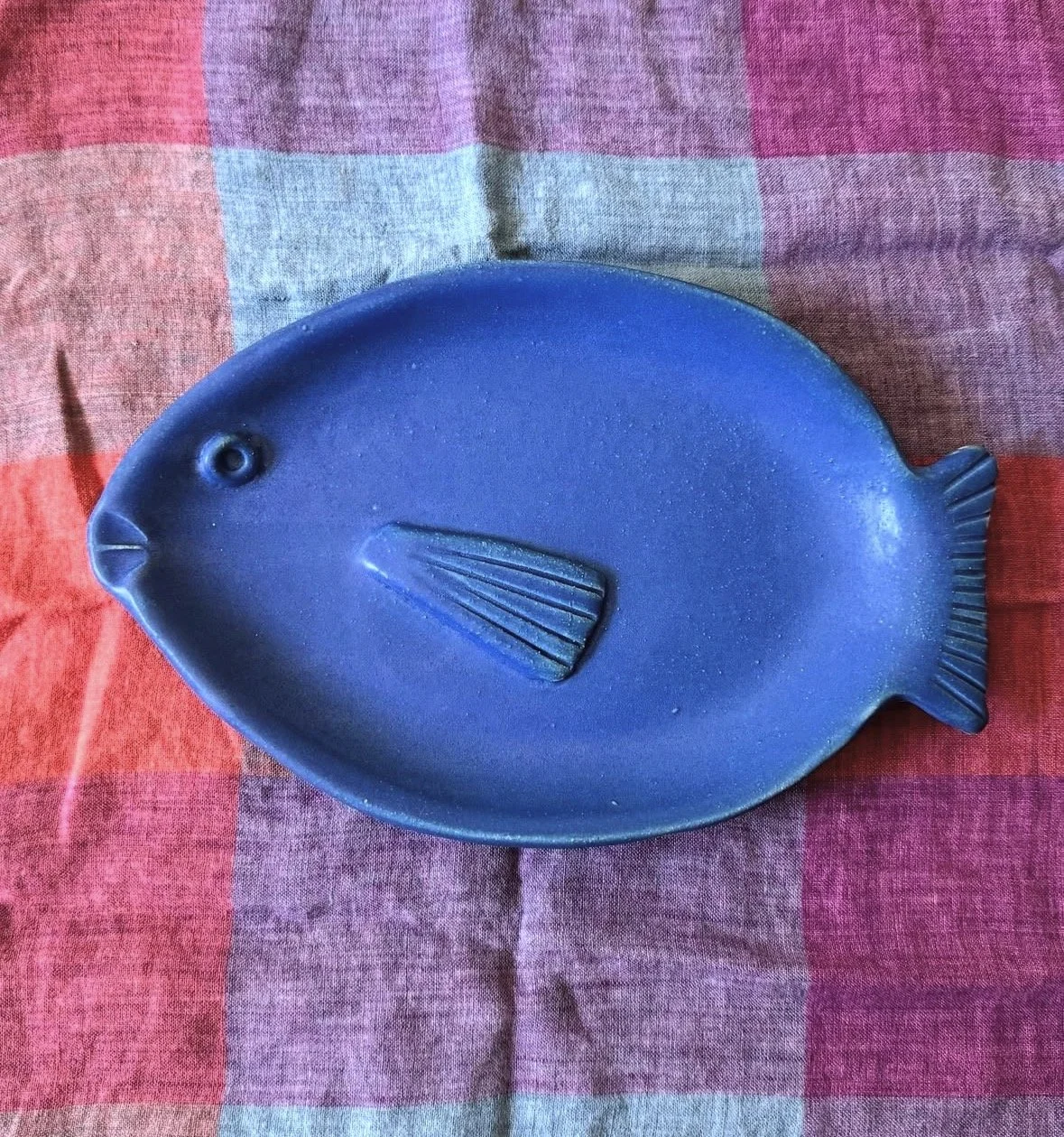 Blue fish-shaped ceramic dish with a prominent fin in the middle, placed on a multicolored fabric background.