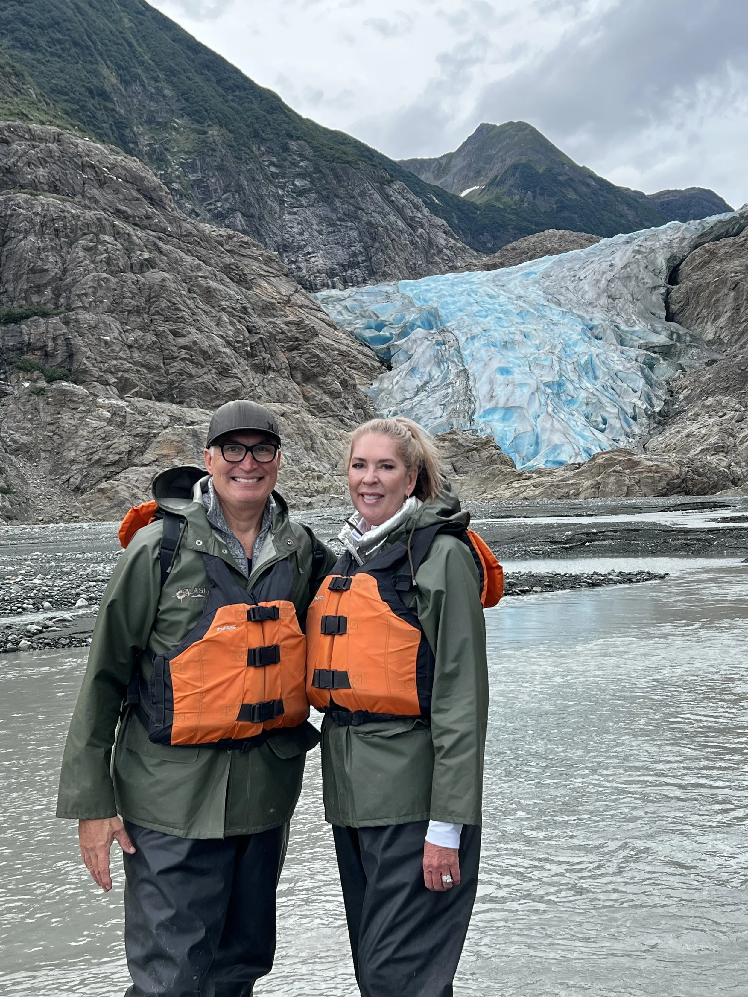 A smiling man and woman in outdoor gear standing in front of a river with a mountain and glacier in the background.