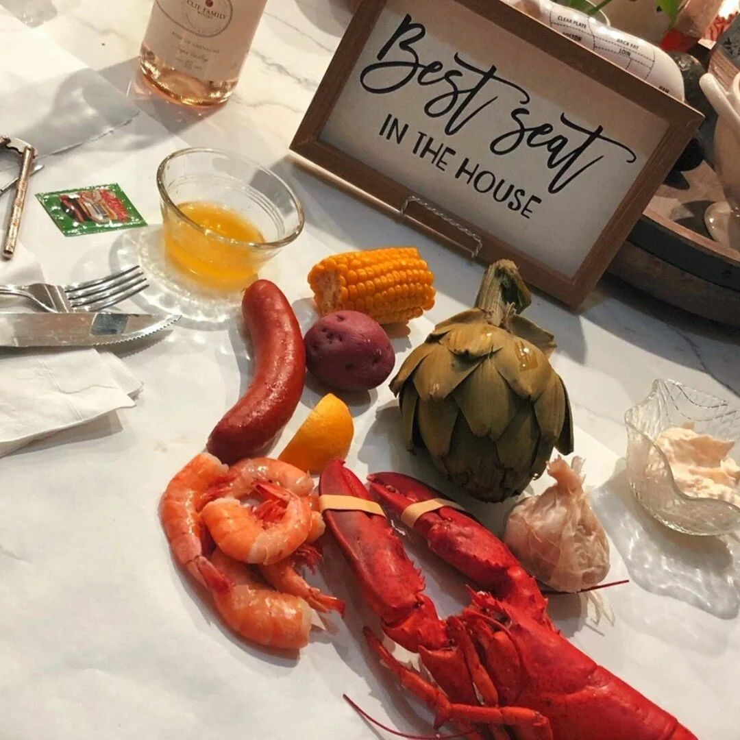 A seafood and vegetable display on a white table with a sign that reads 'Best Seat in the House.' The display includes crawfish, shrimp, a corn on the cob, a potato, a red sausage, a small pumpkin, and a lemon wedge.