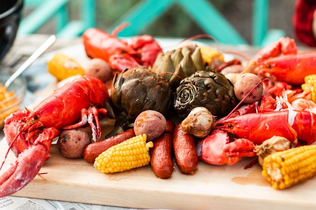 Fresh seafood and vegetables on a wooden cutting board, including lobsters, artichokes, potatoes, carrots, and corn.