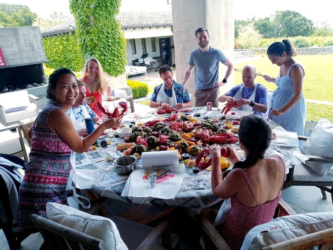 People gathered around a large table outdoors, preparing and eating seafood, including lobsters, on a sunny day.