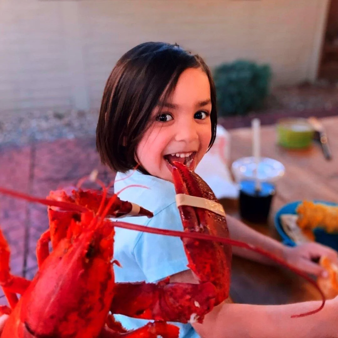 A young girl with short dark hair smiling as she eats a cooked lobster outdoors at a table with various dishes and drinks in the background.