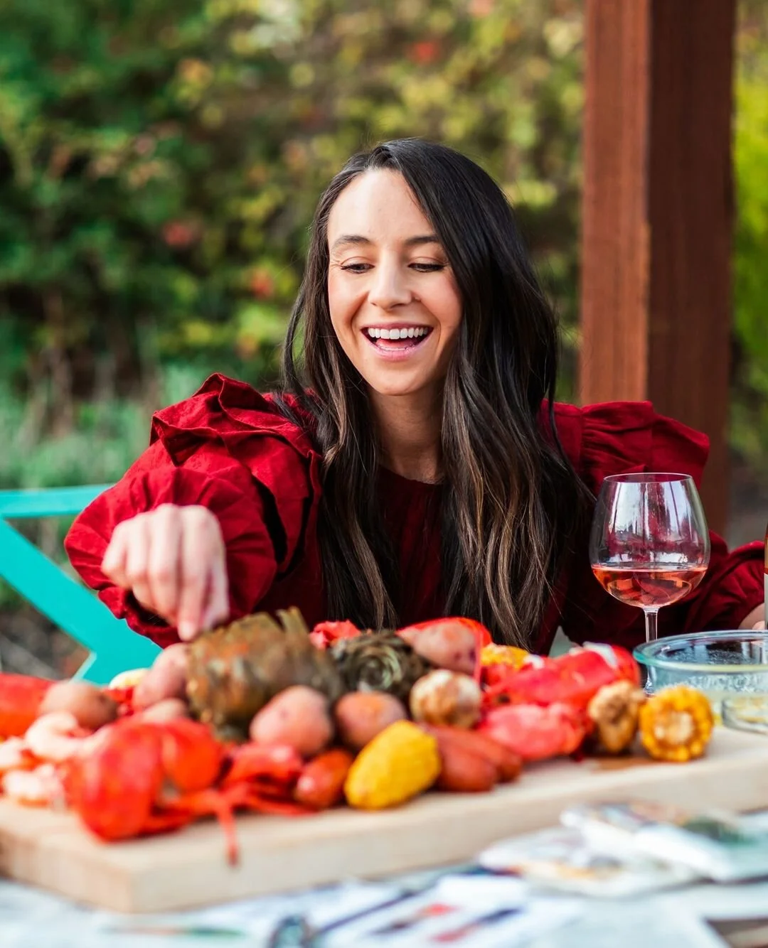 Woman with long dark hair smiling and reaching for food on table outdoors with various vegetables and a glass of rosé wine.