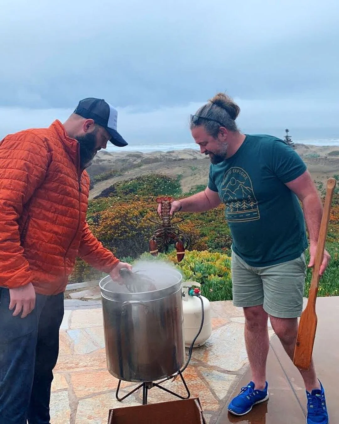 Two men outdoors, one holding a lobster over a steaming pot, the other looking into the pot, with a scenic landscape of bushes, rocks, and an overcast sky in the background.