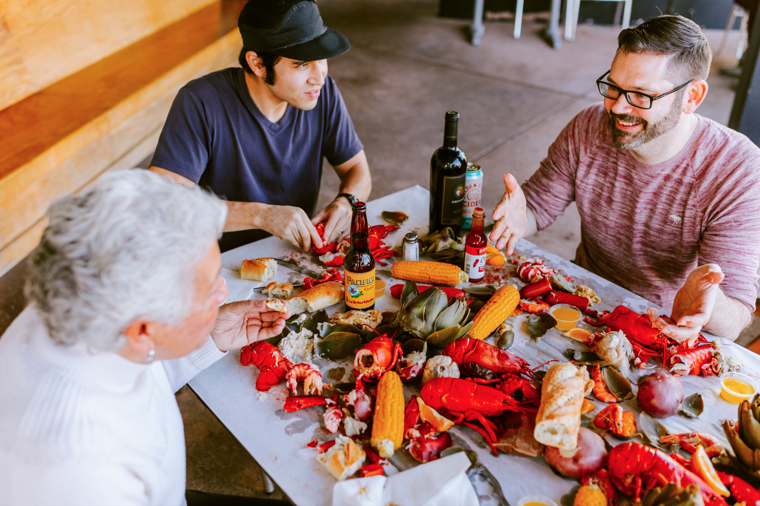 Four people seated at a table with lobster, corn, bread, and drinks, having an outdoor meal.