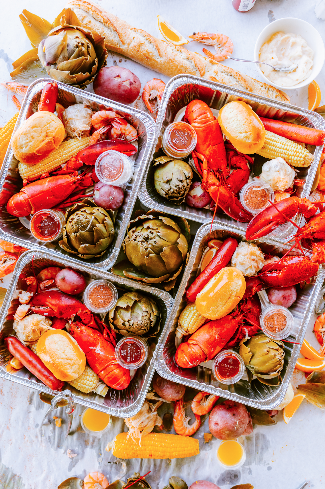 Seafood boils with lobster, crawfish, shrimp, corn on the cob, potatoes, and artichokes in aluminum trays, alongside bread, lemon slices, dipping sauces, and beverages on a white table.