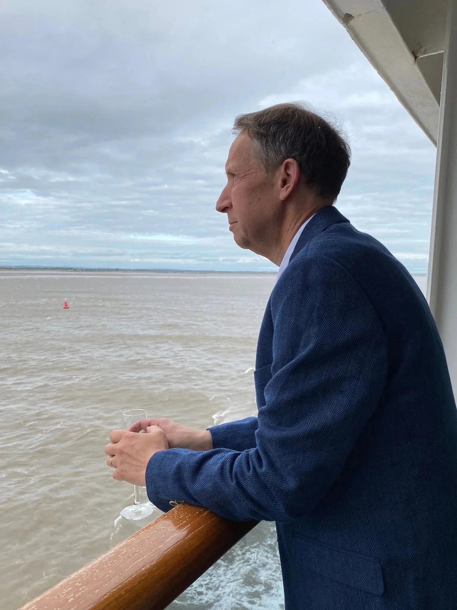 A man in a blue blazer sitting on a boat deck, holding a wine glass, overlooking a cloudy ocean. The boat's wooden railing is visible in the foreground.