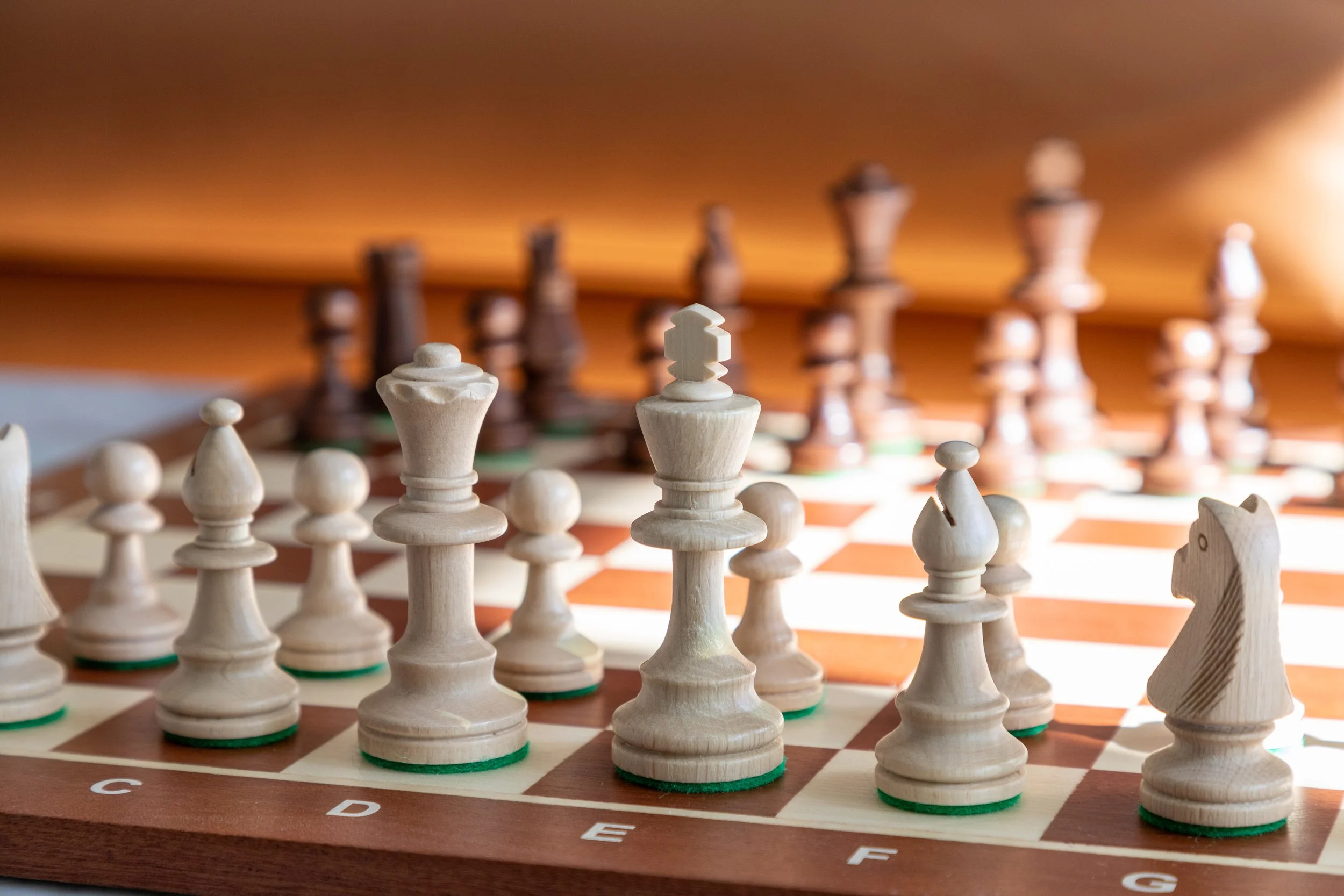 Close-up of a wooden chessboard with white and dark chess pieces arranged for a game, focusing on the white king and queen in the foreground.