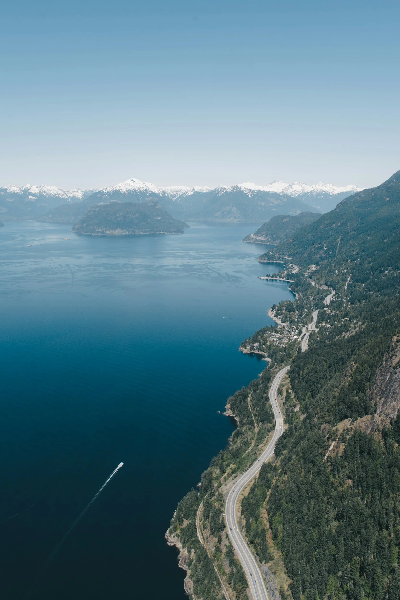 An aerial view of a large body of water with a mountain landscape in the background. There are snow-capped peaks and green forested hills along the shoreline. A winding road runs along the shoreline, and there is a boat creating a white wake on the water.