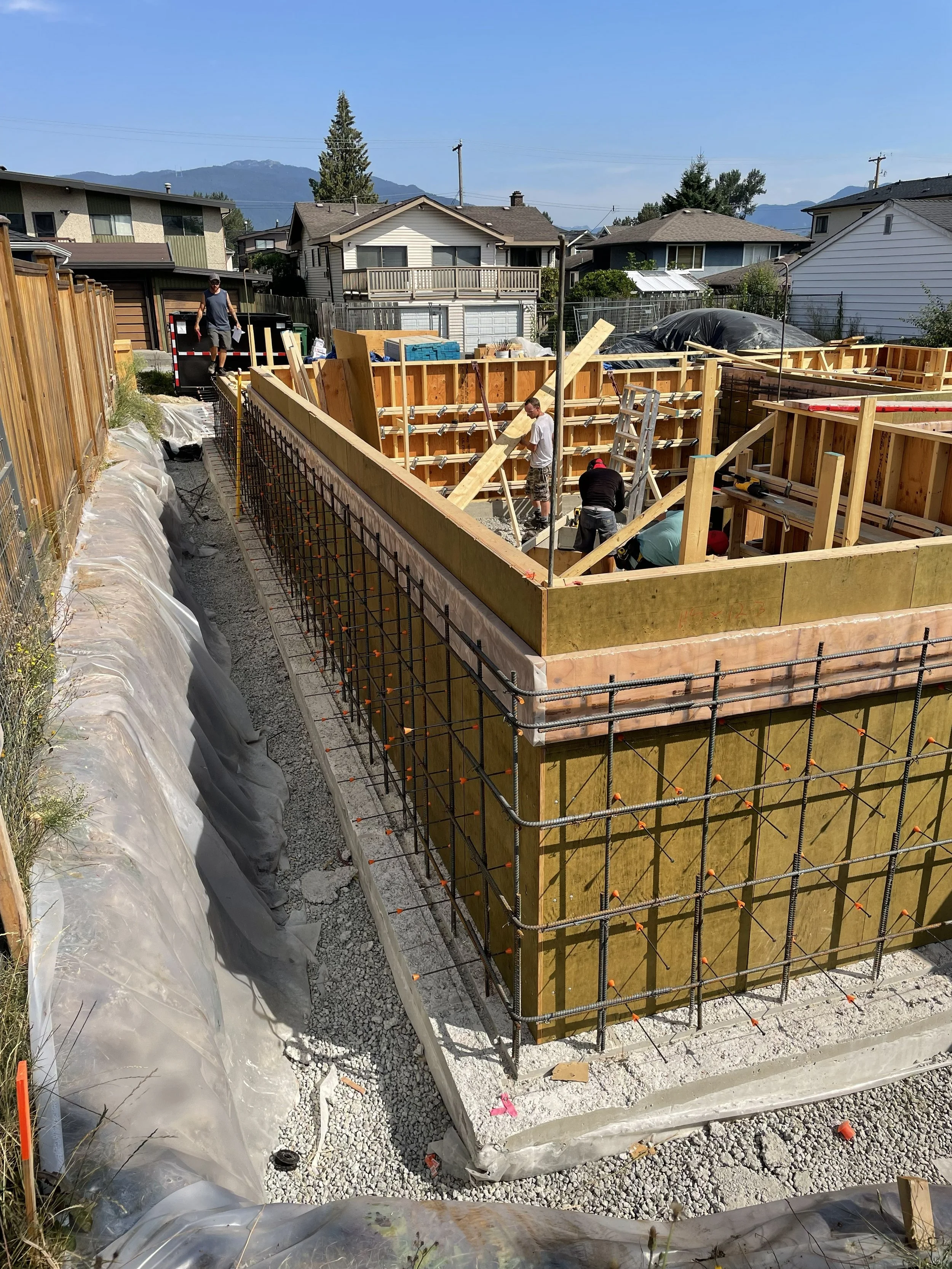 Construction site with wooden framing for a building foundation. Four workers are present, and various construction materials and tools are visible. In the background, various residential houses and mountains can be seen under clear blue skies.