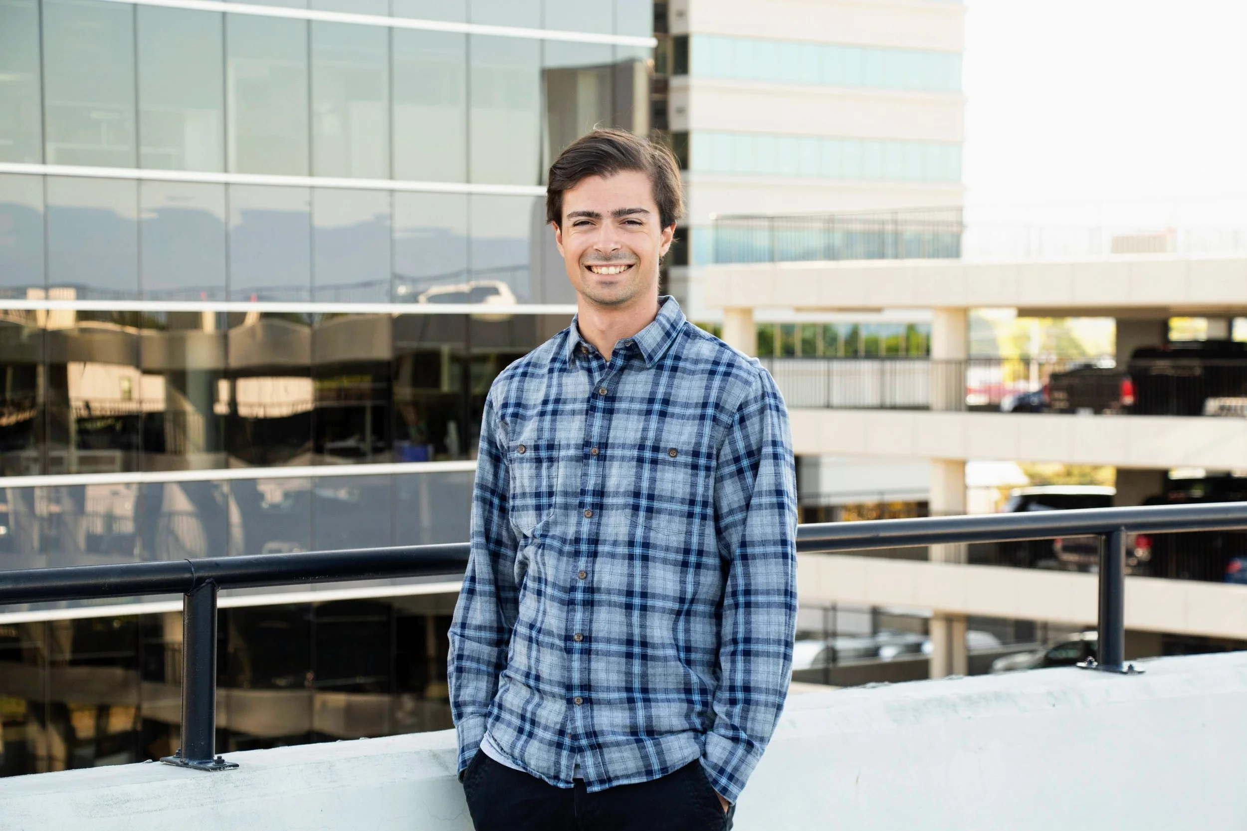 Young man with dark hair smiling standing outdoors in front of a modern glass building wearing a blue plaid shirt with sleeves rolled up and hands in pockets.