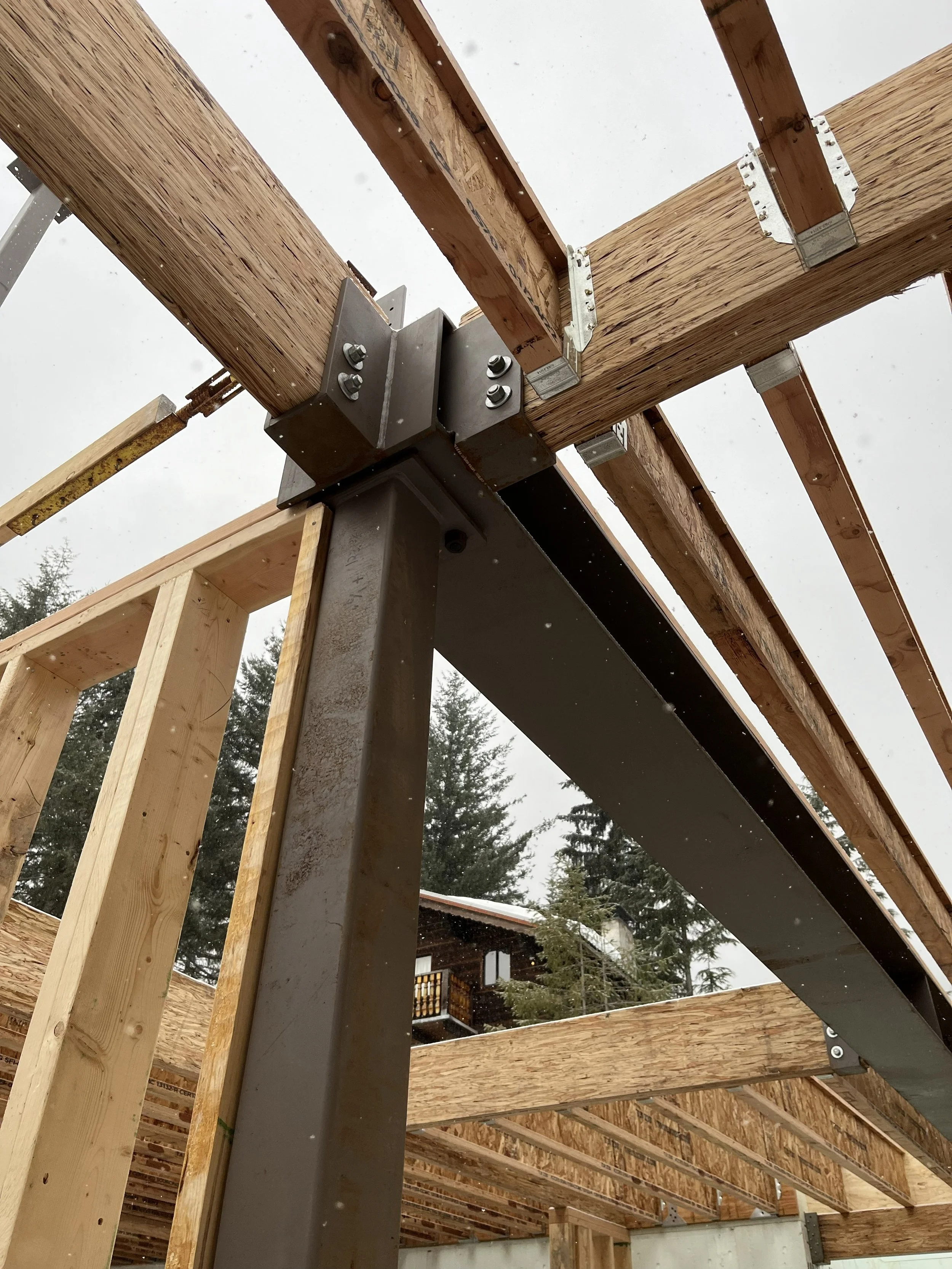 View from the ground looking up at a construction site with wooden beams, metal brackets, and hardware supporting the framework of a building, with trees and a neighboring house visible in the background.