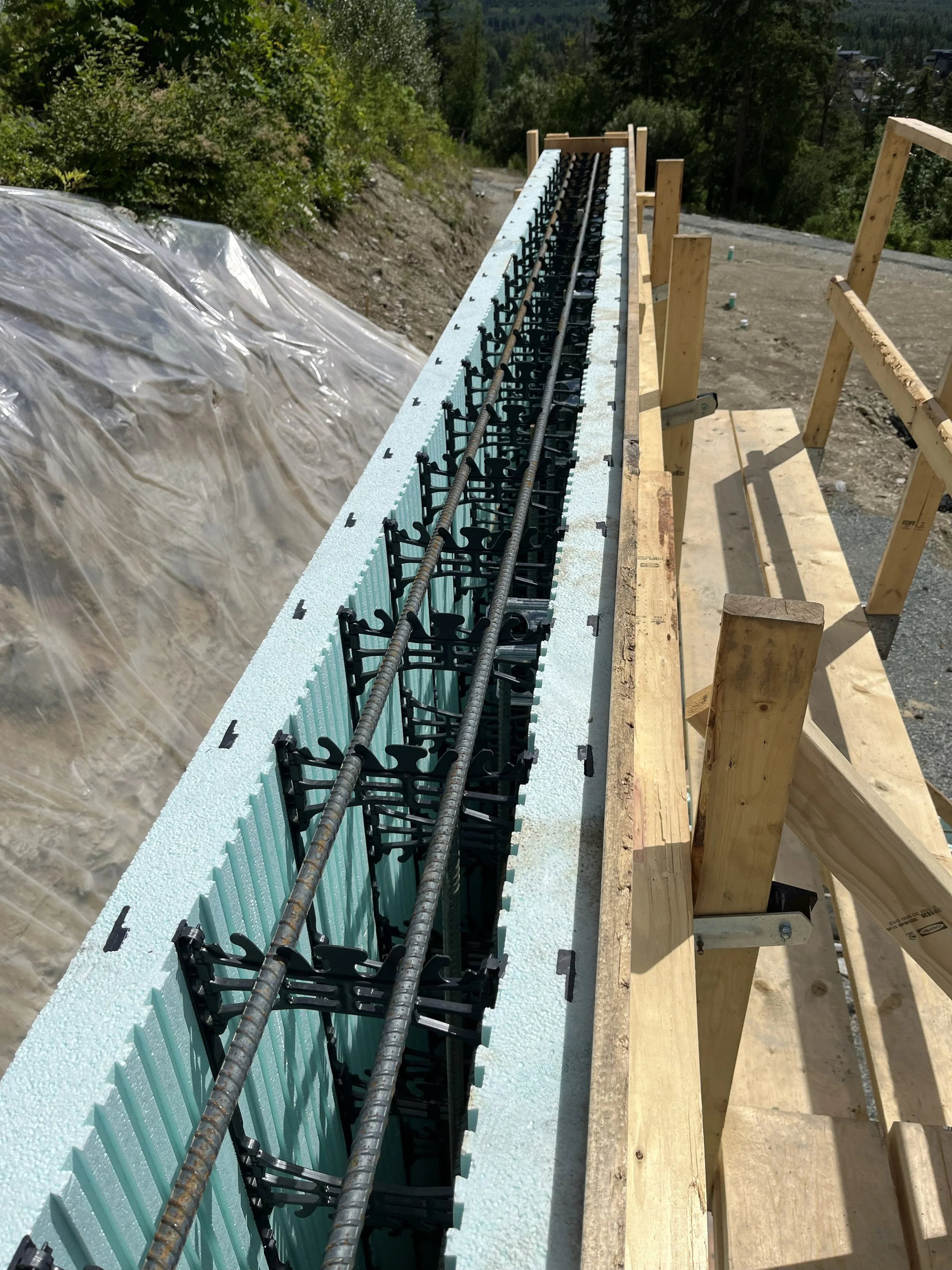 Construction site with wooden framework and foam insulation with rebar on top, situated on a mountainous area with green trees and dirt ground.