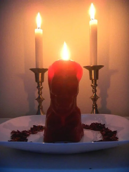 A red candle burning with a flame, positioned in front of two lit candles on candleholders, on a white plate with dark decorative elements, against a plain wall background.