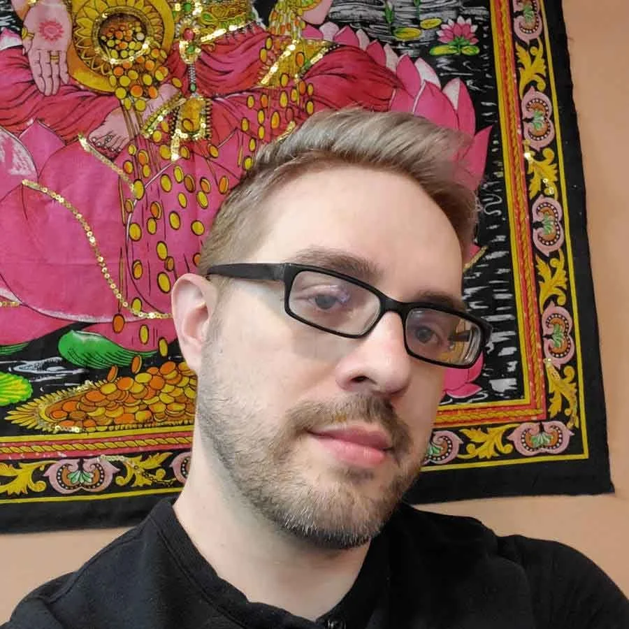 A man with glasses and light brown hair taking a selfie in front of a colorful tapestry featuring a detailed image of a deity or Buddhist figure surrounded by flowers and decorative elements.