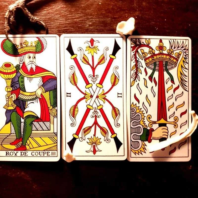 Three tarot cards laid out on a wooden surface. The first card is the King of Clubs, showing a king seated with a crown and scepter. The middle card is the Two of Clubs, displaying crossed club symbols with decorative designs. The third card is the Ace of Clubs, featuring a large stylized club symbol with flames and a hand gripping a sword.