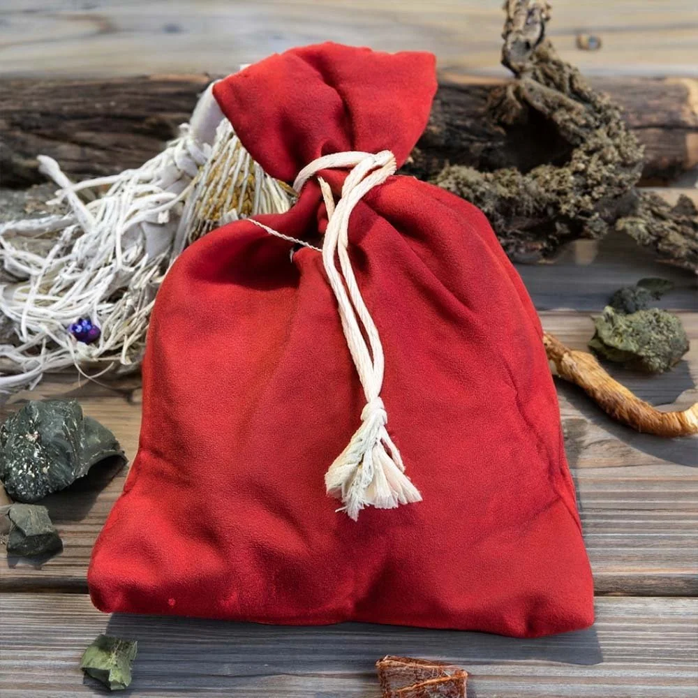 A red cloth bag tied with a white string, placed on a wooden surface outdoors, with natural elements like rocks, driftwood, and moss around it.
