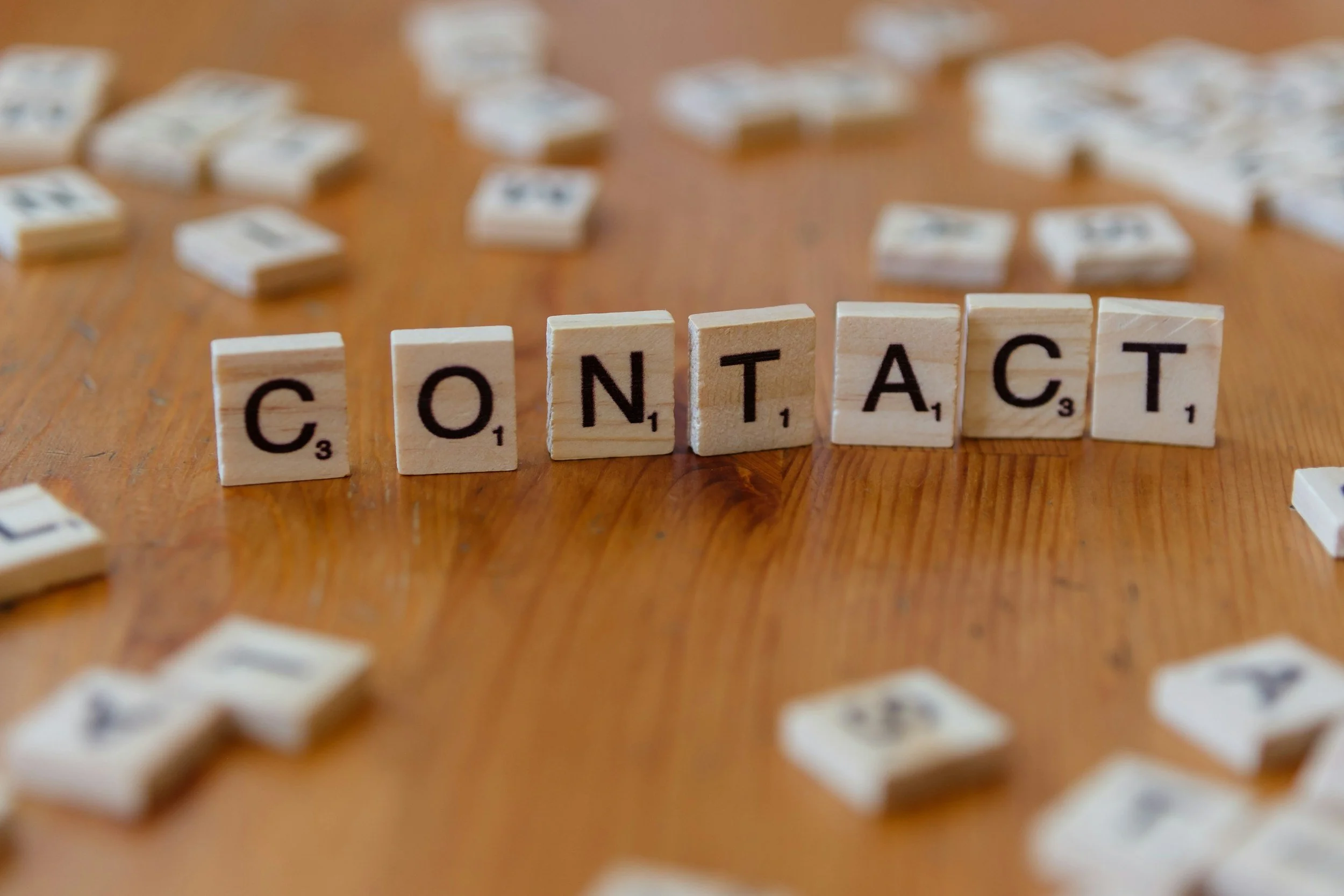 Nahaufnahme von Scrabble-Buchstaben, die das Wort 'CONTACT' auf einem Holztisch bilden, mit mehreren verstreuten Buchstaben in Hintergrund.