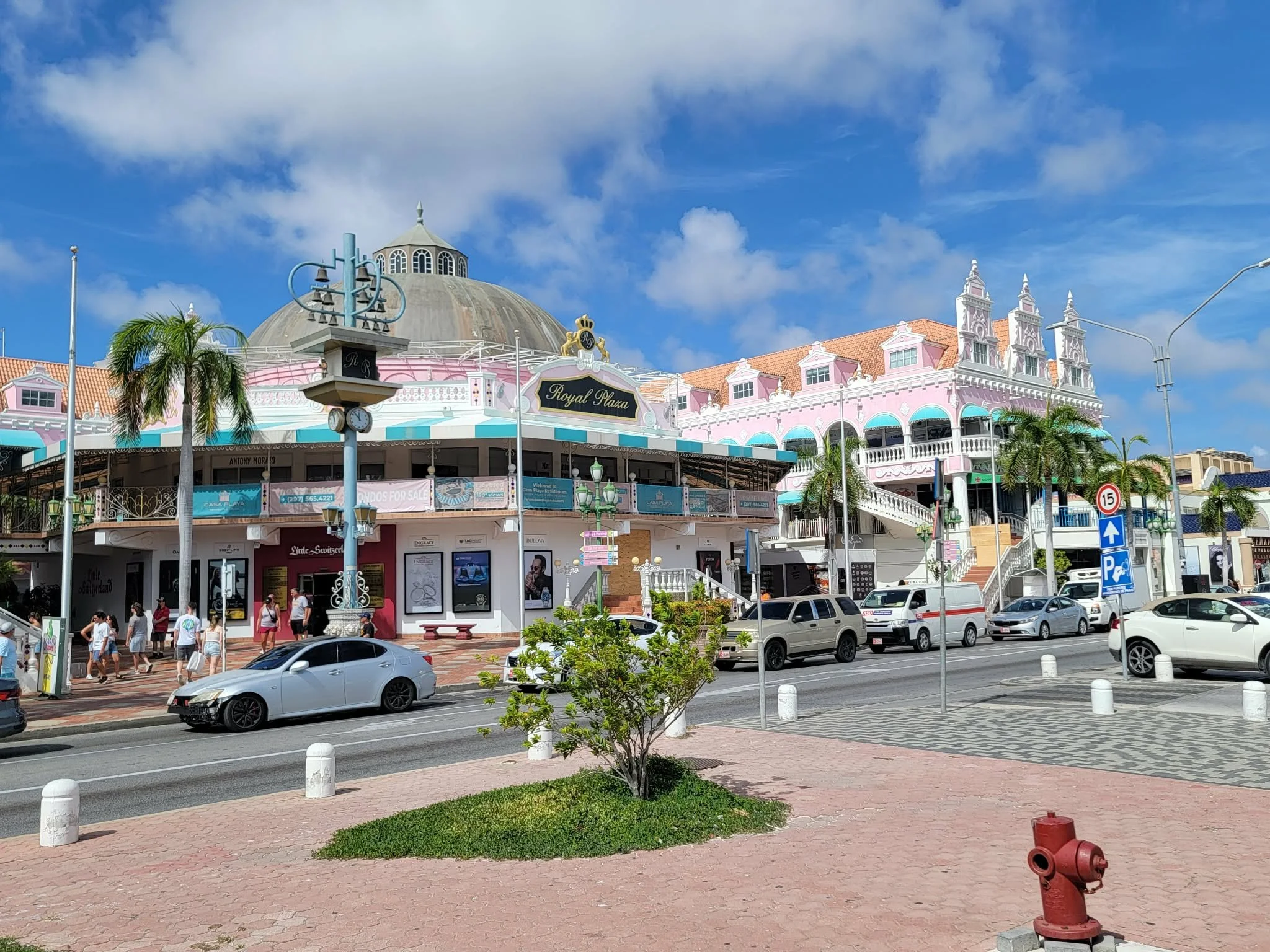 A busy street scene in a tropical location featuring a pink and white building with ornate architectural details, palm trees, and cars parked along the street. There are pedestrians walking on the sidewalk, and a blue sky with some clouds overhead.