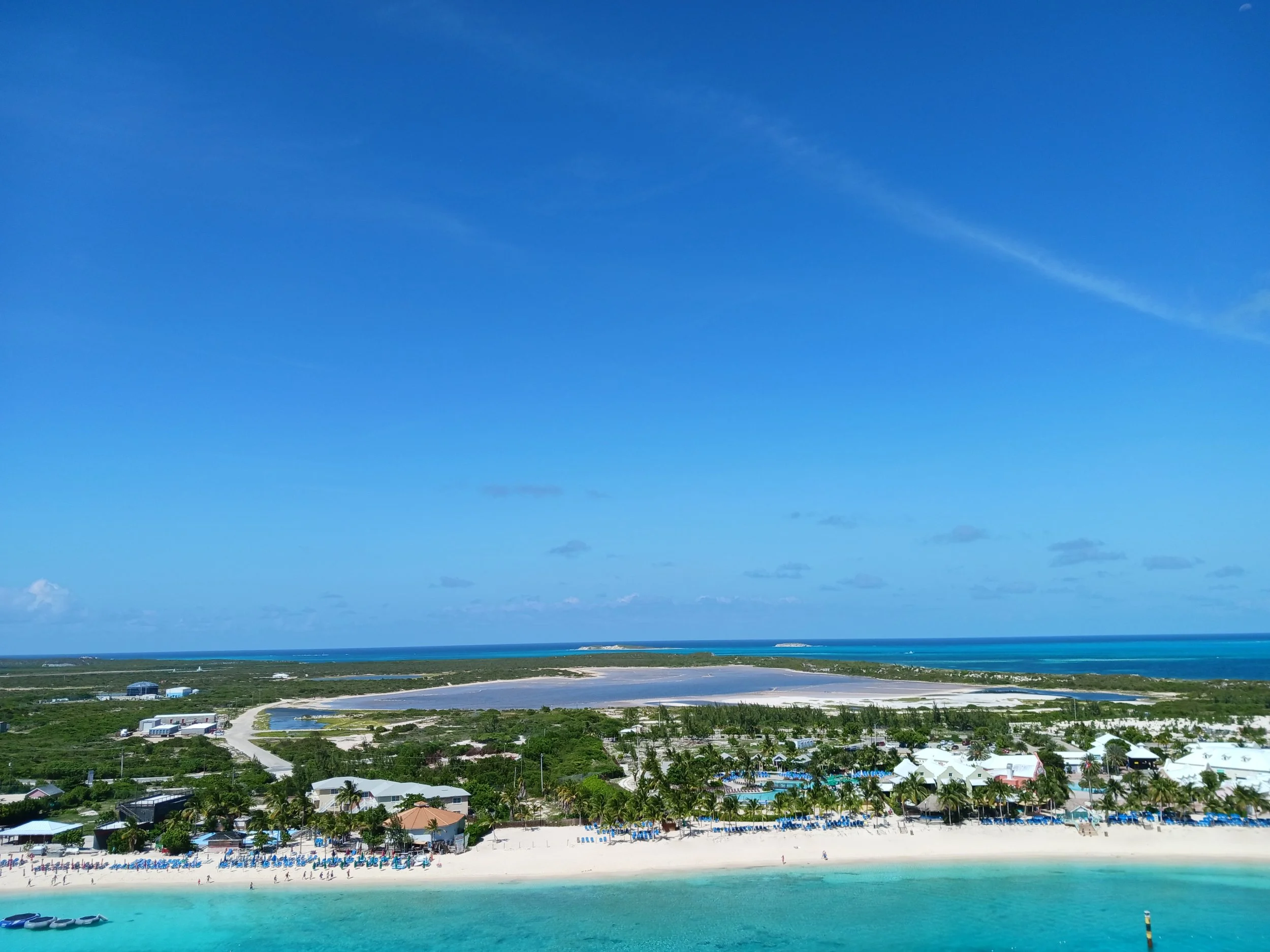 A view of a coastal resort area with a beach, ocean, and buildings, under a bright blue sky with a few clouds.